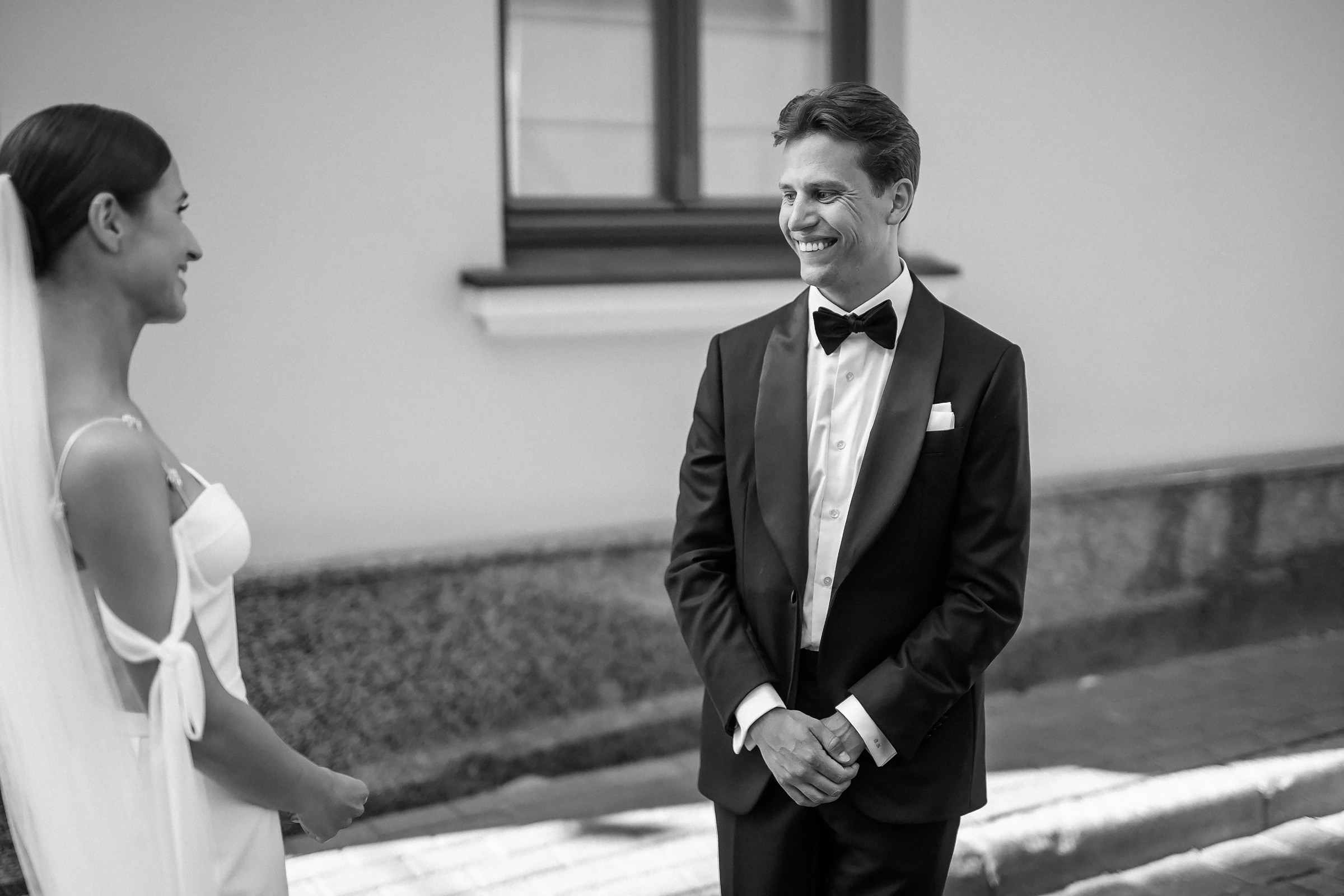 A black and white wedding scene of the bride in a white wedding dress with a veil and a groom in a black tuxedo with a bow tie standing opposite each other. They are in front of a building with a window and a sidewalk.