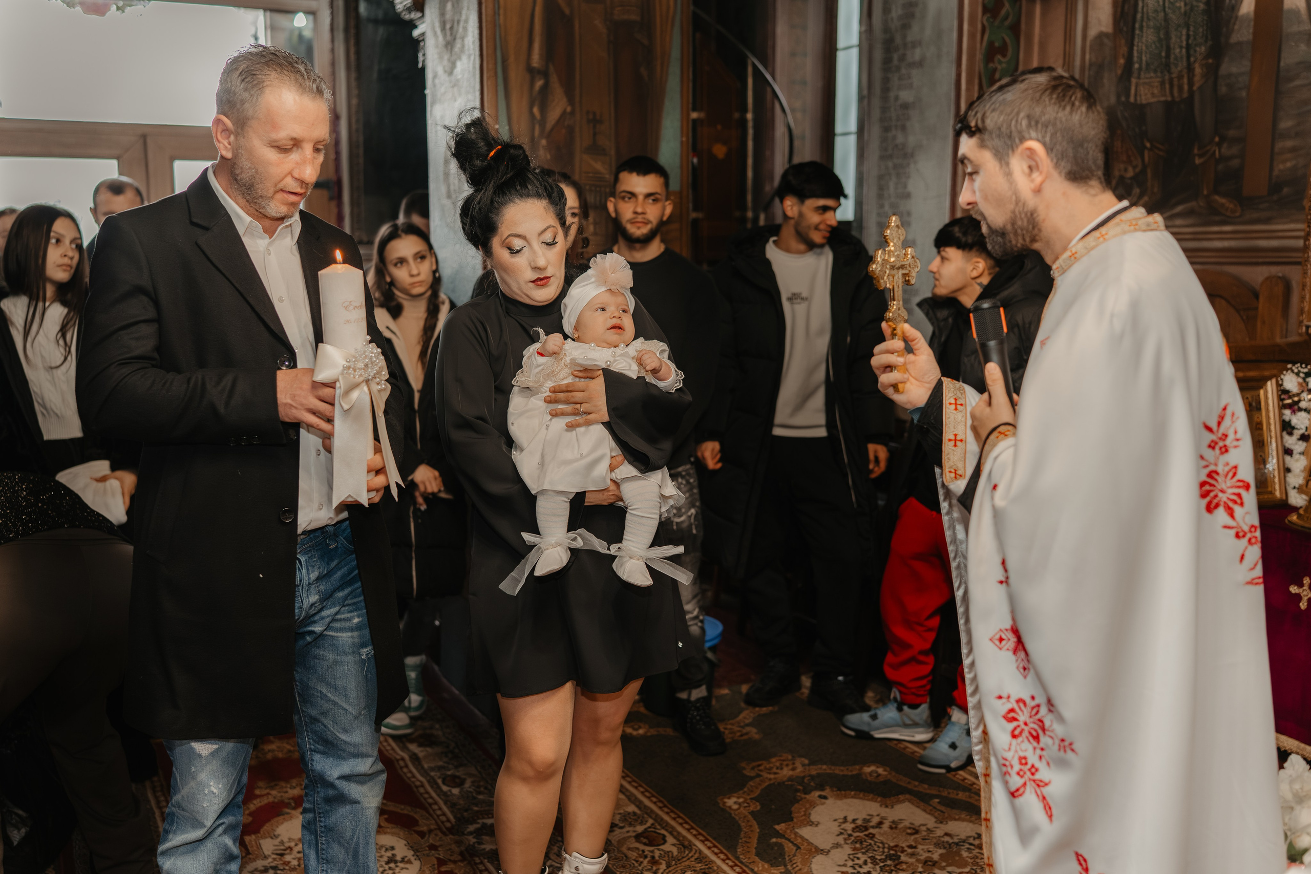 Preotul face o binecuvântare finală pruncului Eveline Maria, acum îmbrăcat în haine noi, ținând crucea în mână deasupra ei. Nașii și familia asistă solemn la acest ultim act al ceremoniei de botez.