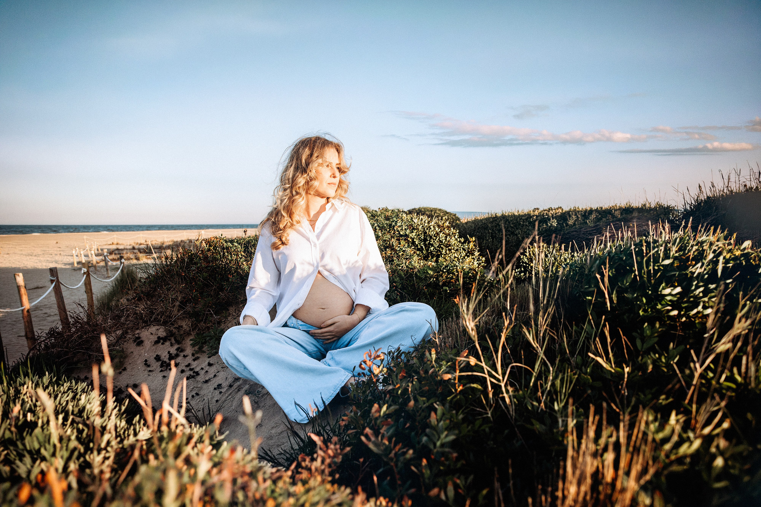 Mujer embarazada sentada con las piernas cruzadas sobre vegetación costera en Valencia, España, sosteniendo suavemente su barriga durante una sesión de fotos de embarazo al aire libre con cálida luz del atardecer y paisaje mediterráneo.