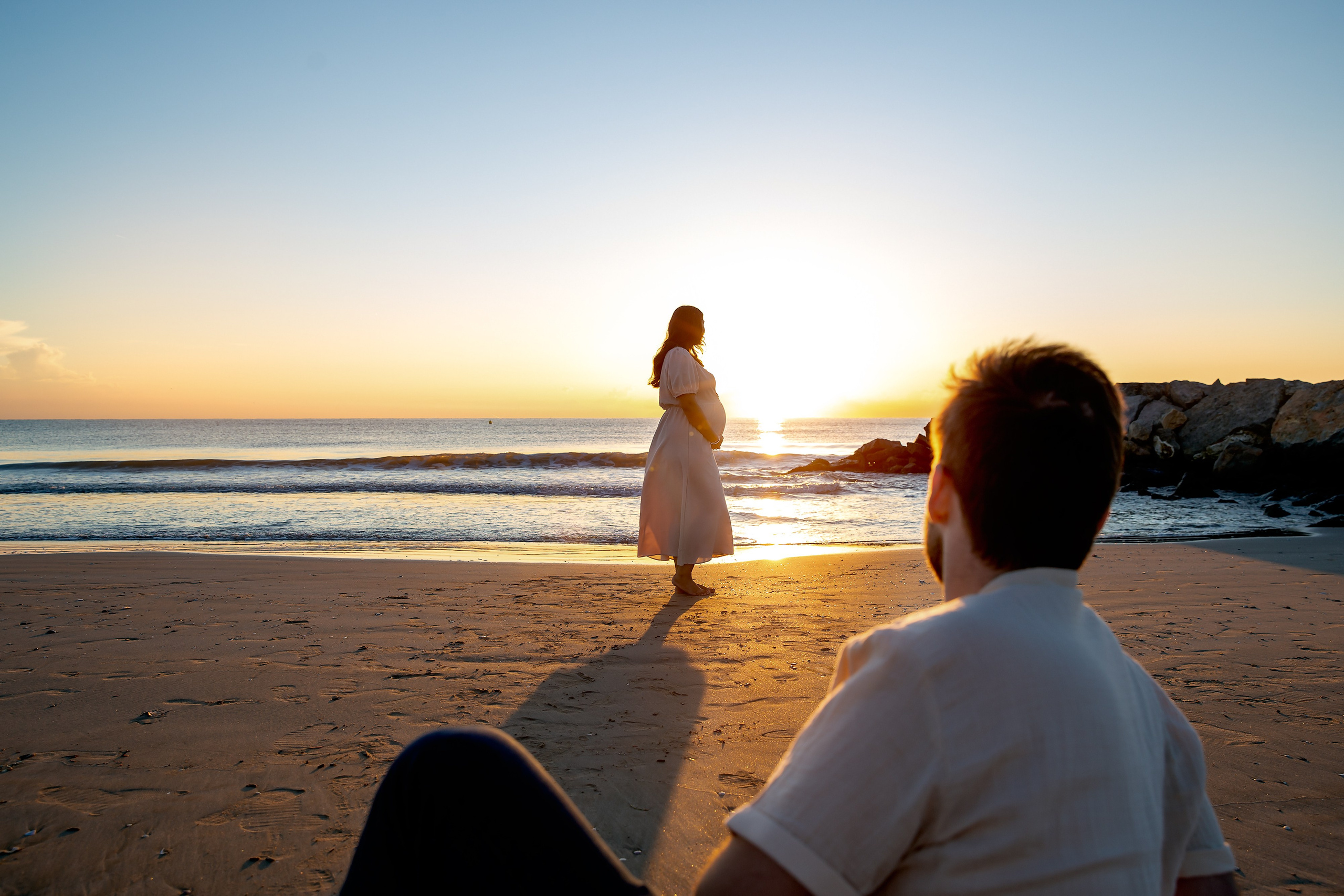 Sesión de fotos de maternidad al atardecer en la playa de Valencia, España, que captura un momento sereno mientras una mujer embarazada se encuentra iluminada por la luz dorada y su pareja la observa con ternura — perfecta para quienes buscan sesiones de fotos de embarazo emocionales y naturales en Valencia y en toda España.