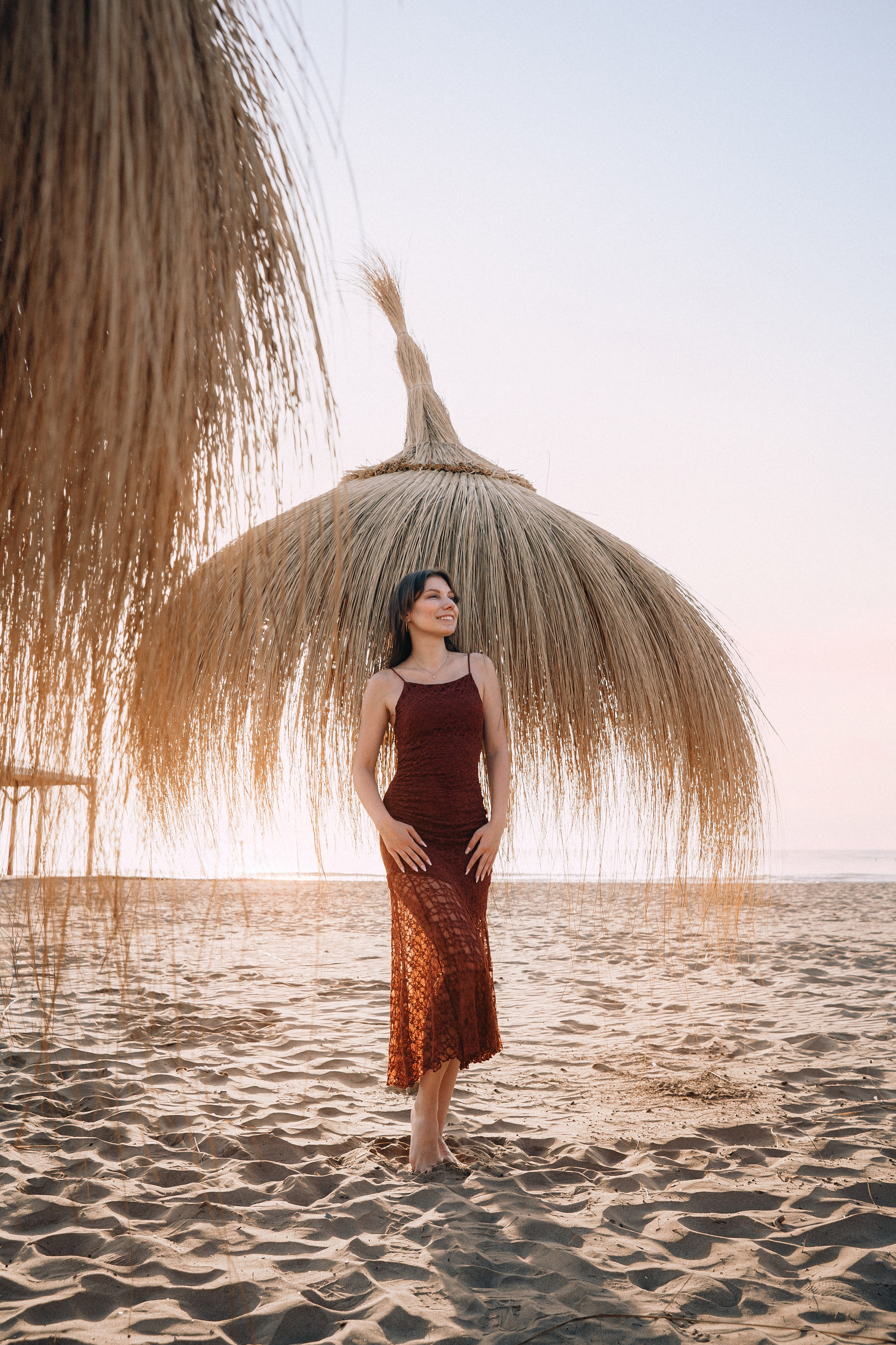 Пляжный портрет на закате: женщина в платье цвета ржавчины стоит под соломенным зонтом в Кальпе, Испания — сочетание бохо-элегантности и природной красоты побережья, идеально подходит для расслабленных и художественных портретных фотосессий в Кальпе и по всей Испании.