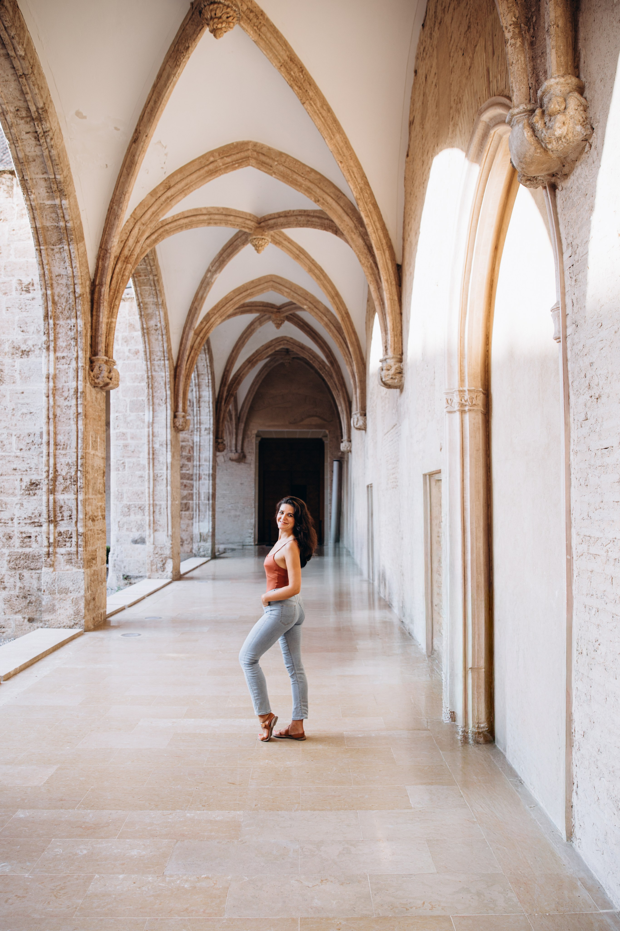 Женская фотосессия в Валенсии, Испания, с уверенной женщиной в историческом готическом каменном коридоре с изящными арками и мягким естественным светом, создающим стильную художественную атмосферу — идеально для женской фотографии, портретных сессий, лайфстайл-съёмок, тревел-портретов и профессиональных женских фотосессий в Валенсии и по всей Испании.