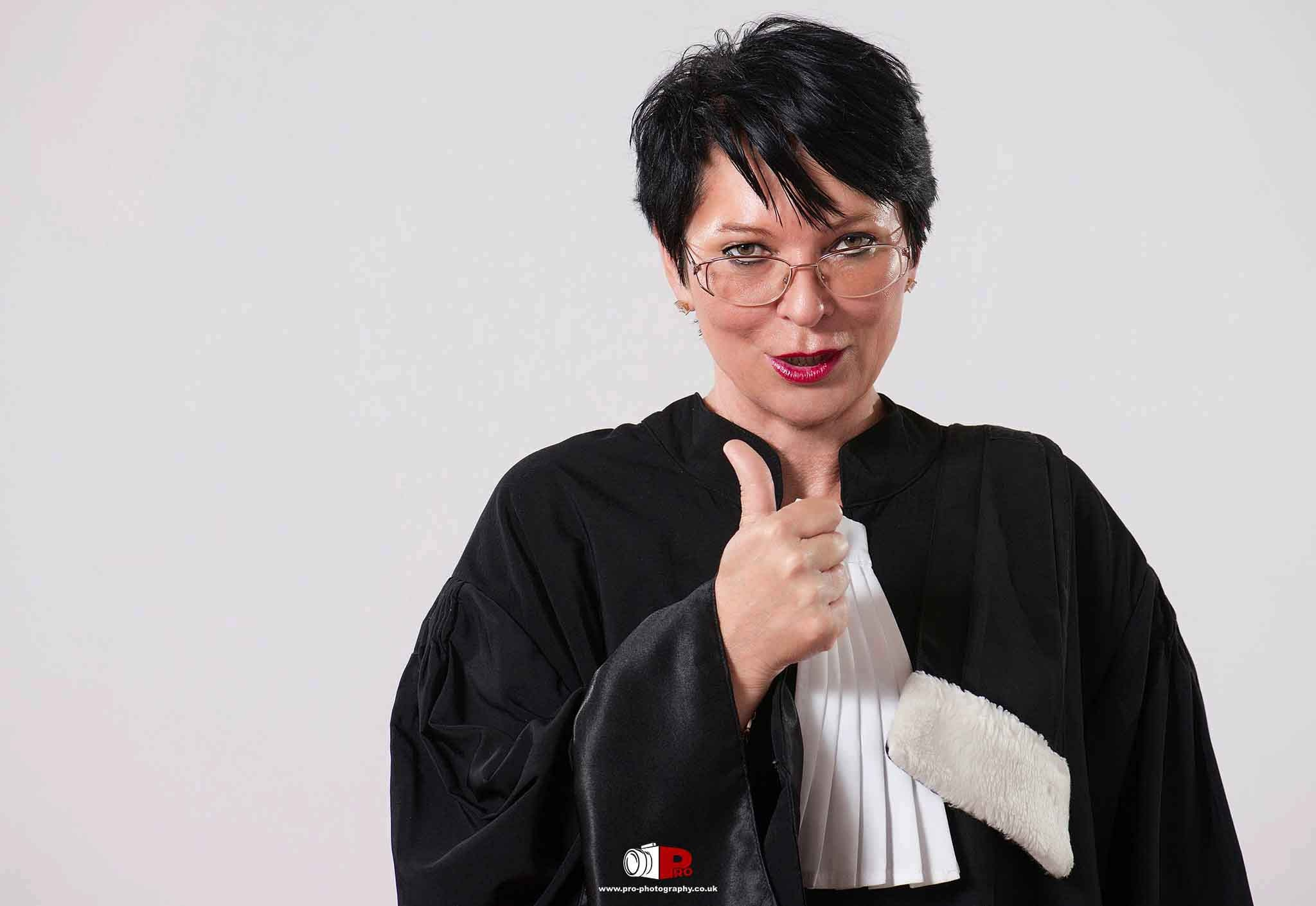 Professional portrait of a female lawyer in traditional court robes giving a thumbs-up gesture with a confident smile.