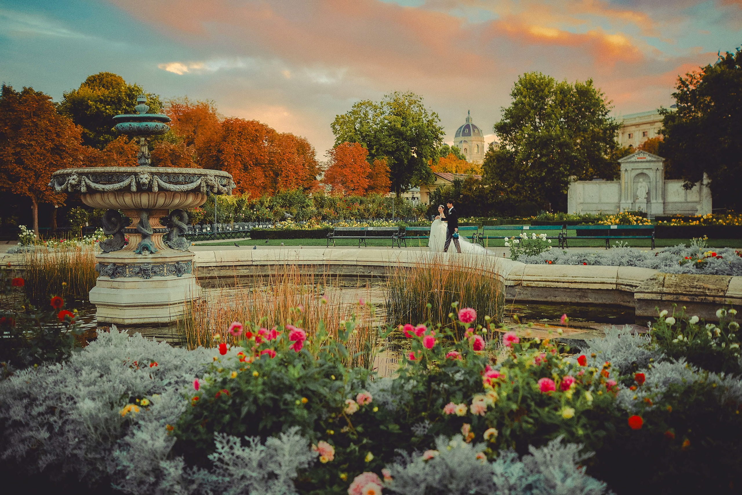 Young Japanese couple in wedding attire walking in sunset-lit autumn Volksgarten, Vienna.