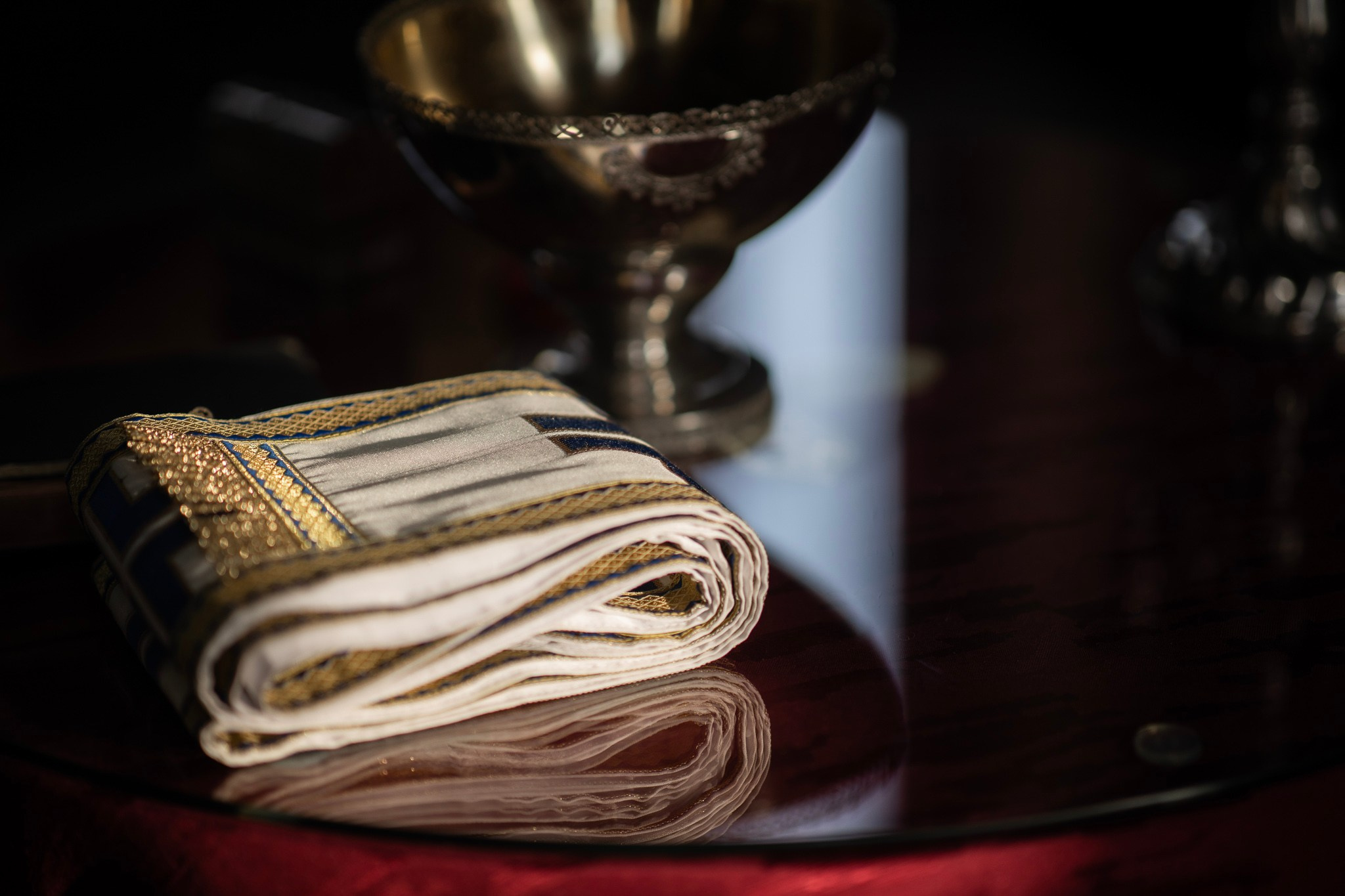 Presavijeni jevrejski tallit (molitveni šal) sa zlatnim obrubom na reflektujućem tamnom stolu, sa zamućenom srebrnom čašom u pozadini.