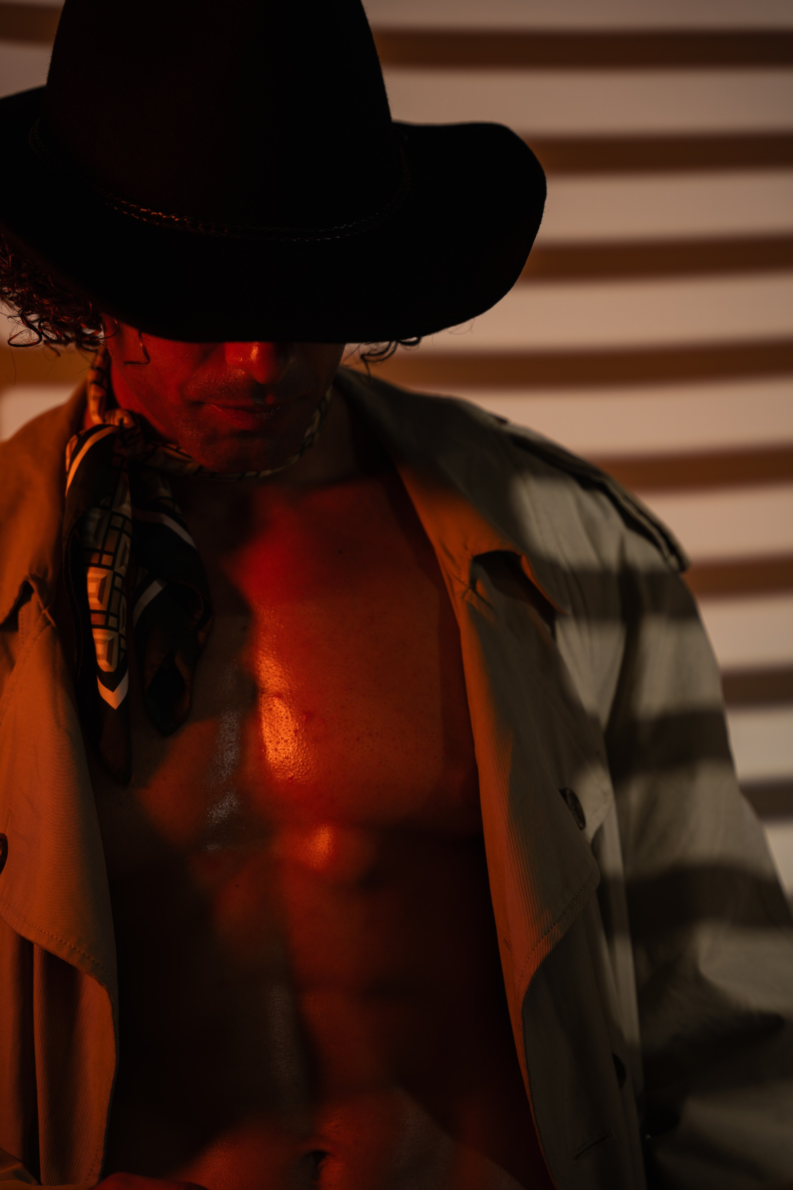 Concept portrait of a figure wearing a hat and open shirt, photographed in warm striped light with a dark cinematic editorial mood.