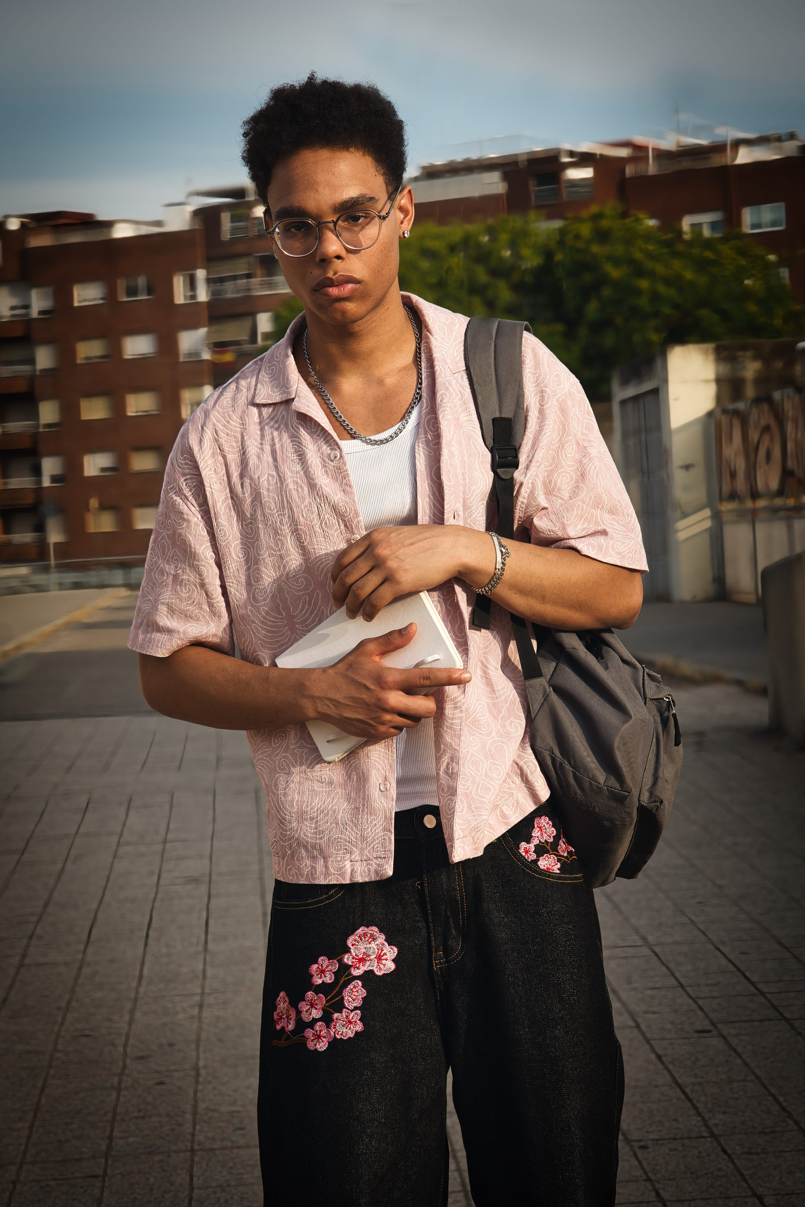 Posa con actitud luciendo un outfit de streetwear casual para hombre, con gafas, camisa estampada y mochila.