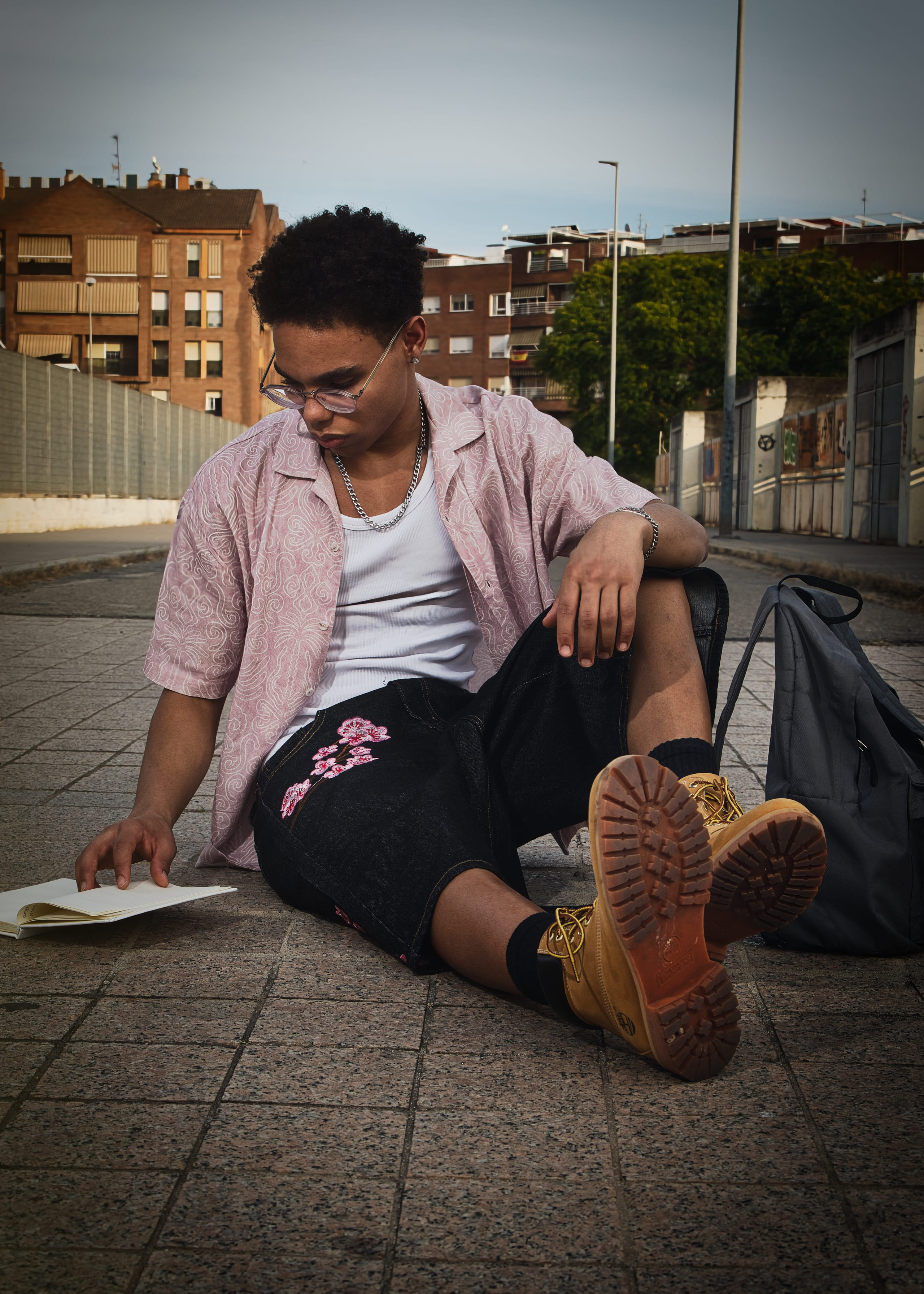 Hombre joven sentado en el suelo, con gafas y botas, disfruta de un momento de lectura con su mochila al lado.