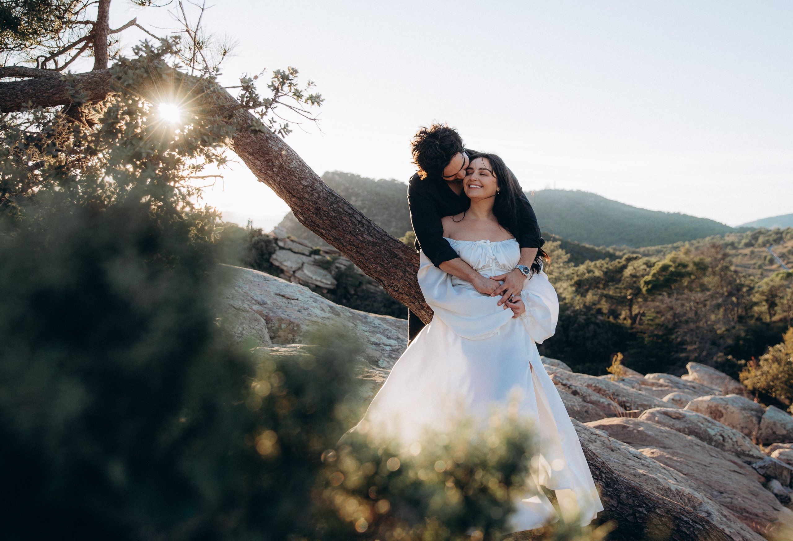 Bride and groom embracing near a windswept tree during their intimate mountain elopement in Barcelona, Spain, surrounded by golden sunset light. The romantic destination wedding portrait highlights the natural landscape and cinematic atmosphere.