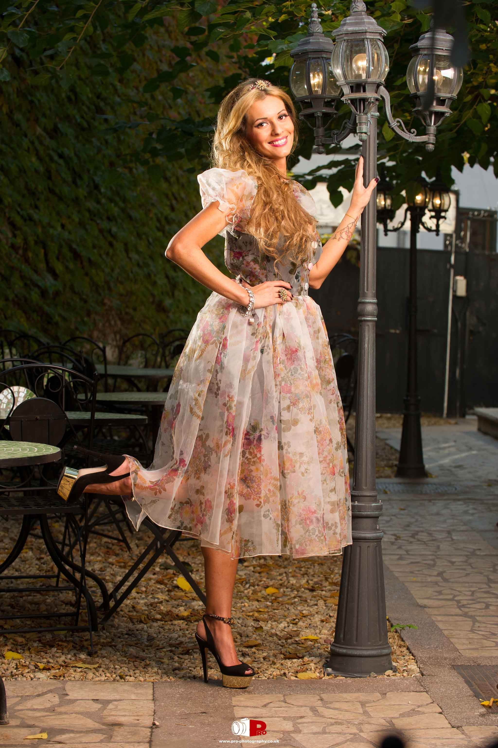 A smiling woman in a floral vintage dress posing playfully beside a decorative lamp post in an outdoor setting.