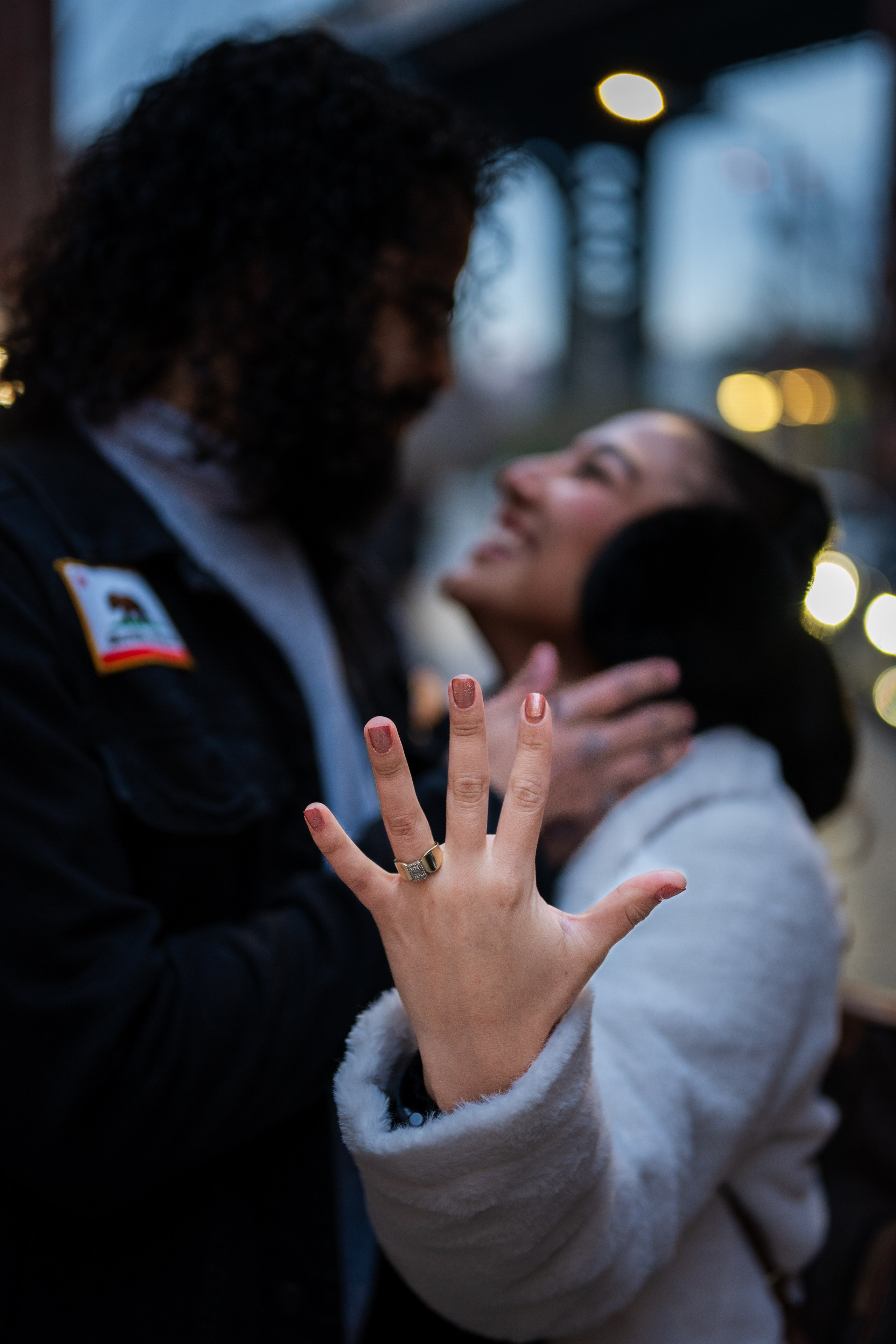 Close-up couple moment in DUMBO at night with focus on a hand showing a ring, romantic NYC engagement-style portrait.
