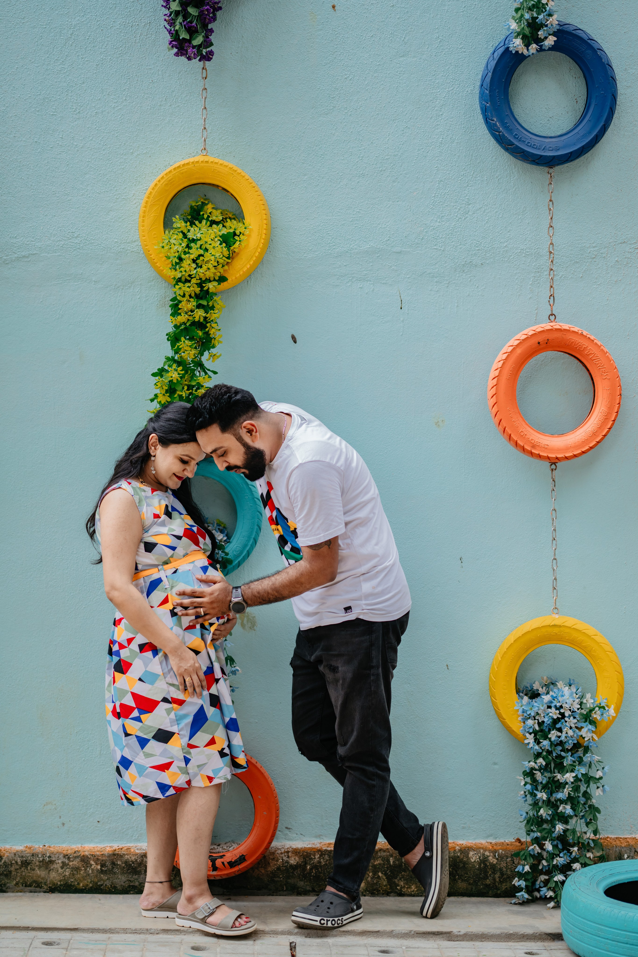 Outdoor maternity photoshoot in Bengaluru featuring a man in a white graphic t-shirt and a woman in a geometric print dress posing against a blue wall with colorful painted tires.