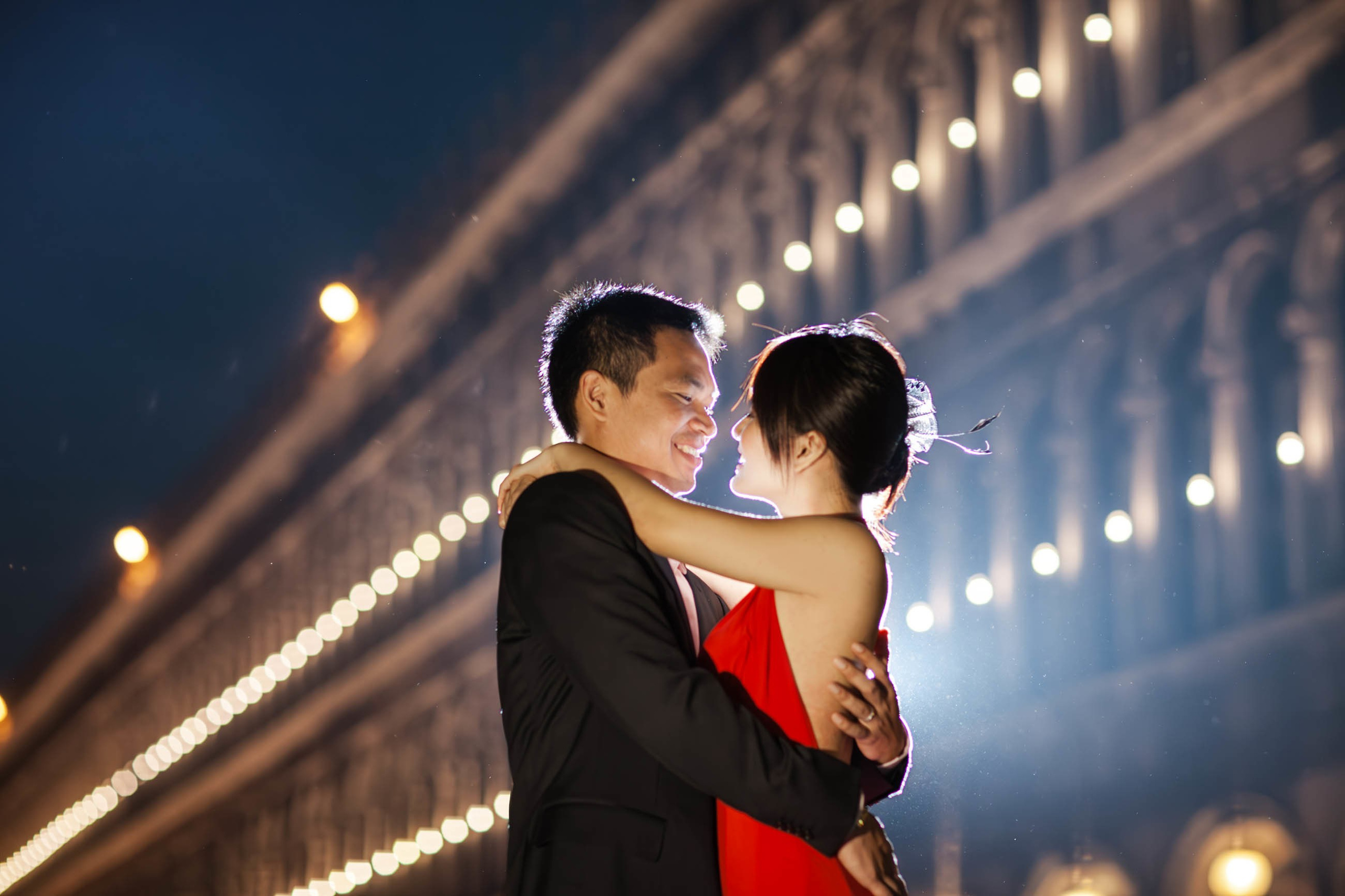 Smiling Thai woman in sexy red dress embraced by fiance for night portrait at San Marco Venice.