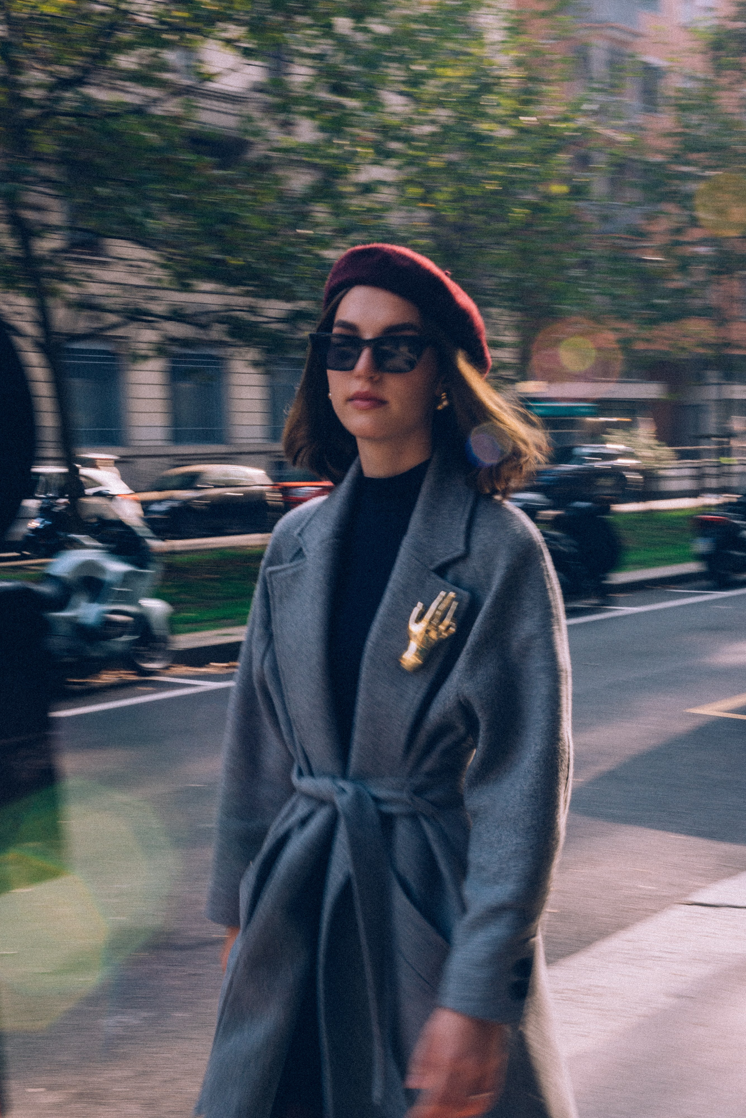 Effortless Milan street style: a woman in a gray coat and burgundy beret walks along a tree-lined avenue, blending fashion with city charm