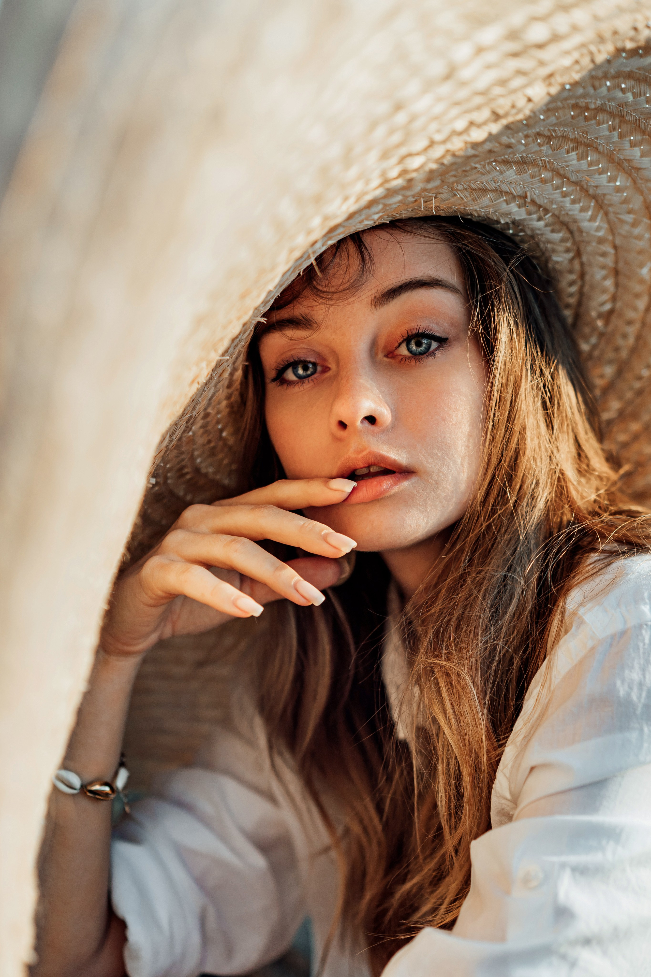 The model, wearing a white bikini and a sheer cover-up, sits under a wide-brimmed hat, lost in thought. The soft light filtering through the hat creates a play of shadows on her face
