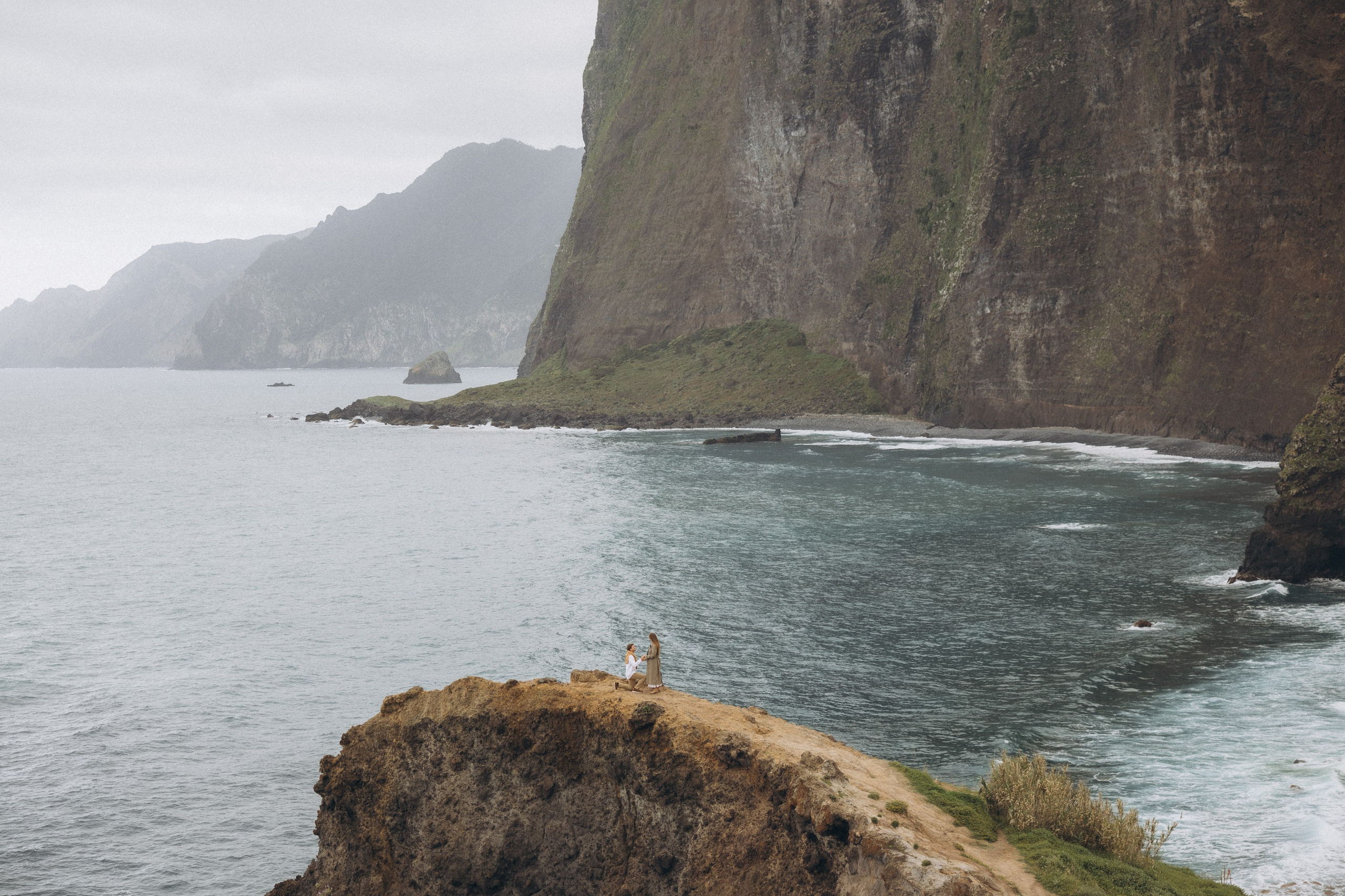 Beautiful engagement moment by the ocean in Madeira, Portugal, as one partner kneels to propose while waves crash in the background