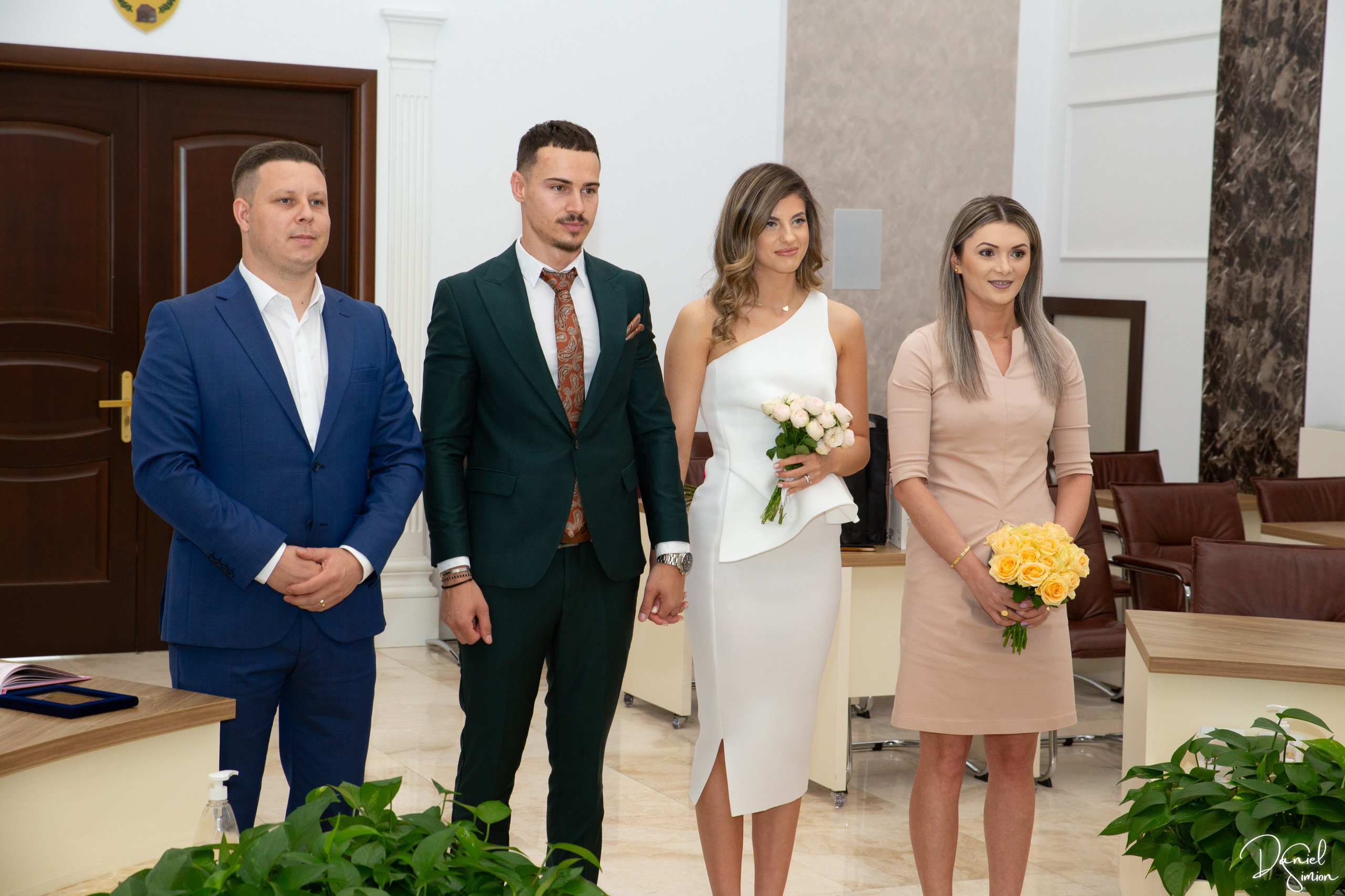 Couple and two attendants standing indoors holding bouquets in a formal setting.