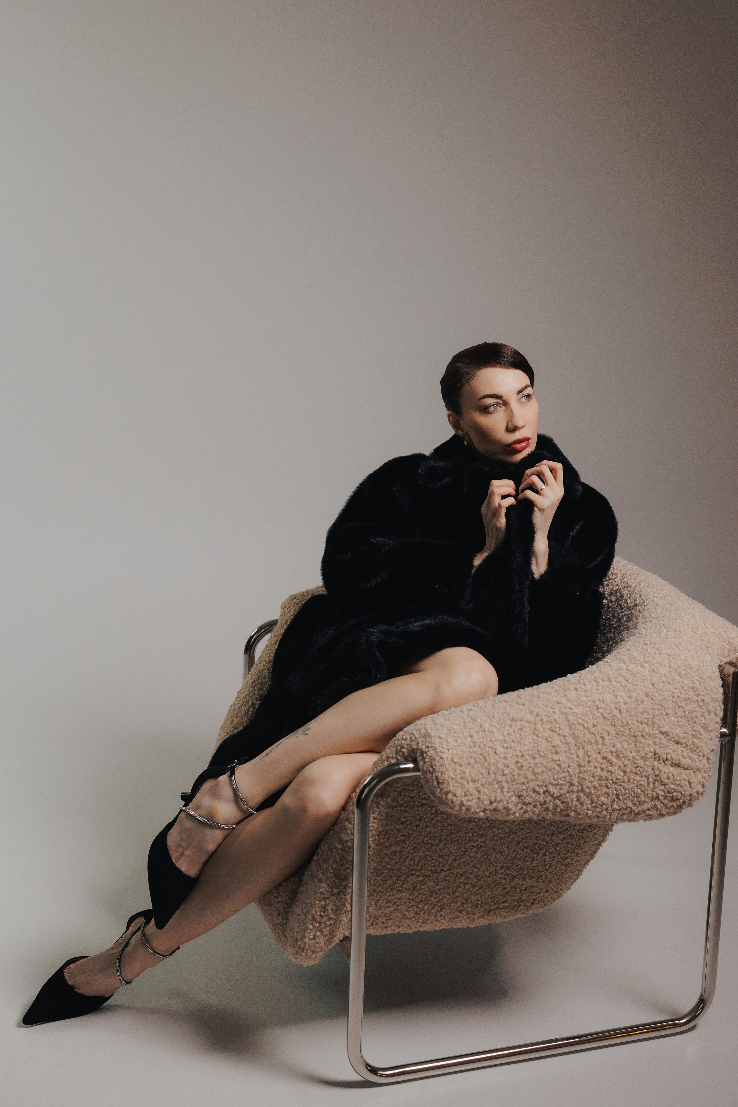 Minimalist studio portrait of a woman in a black fur coat sitting in a chair with dramatic shadows and cinematic lighting, photographed in Leipzig, Germany. Минималистичный студийный портрет девушки в чёрной шубе, сидящей в кресле с драматичными тенями и кинематографичным светом, снятый в Лейпциге, Германия.
