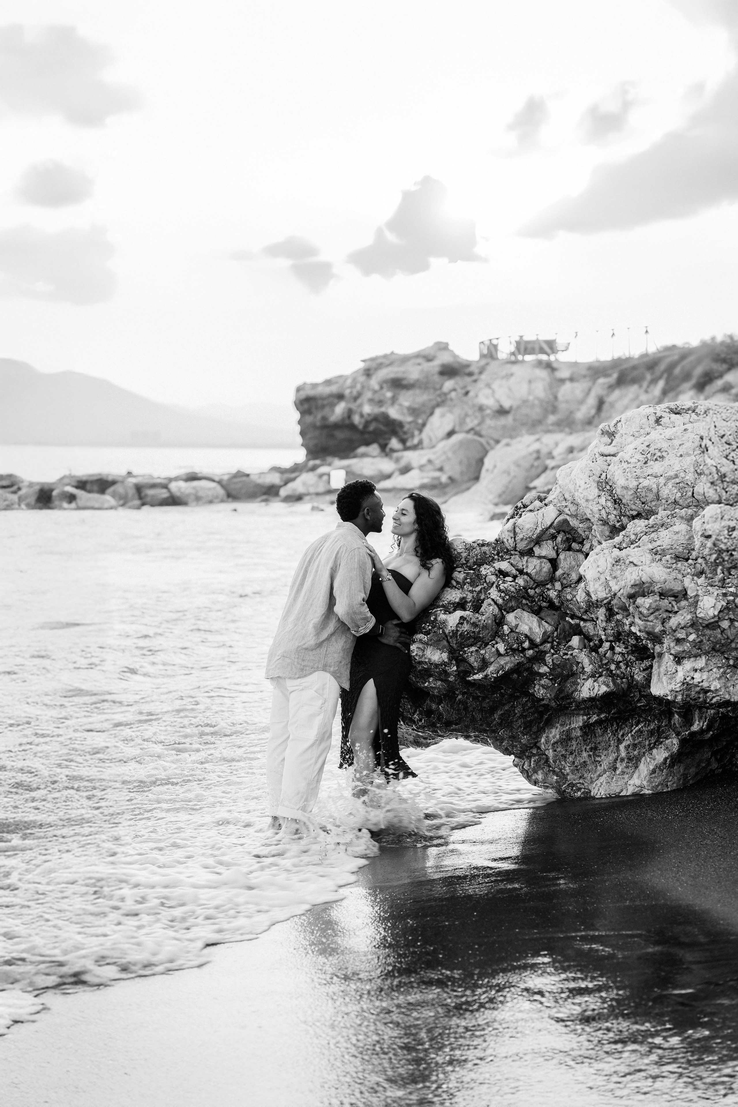 Romantic destination engagement moment in Málaga as the couple sits together by coastal rocks after a seaside proposal. Black-and-white proposal photography capturing intimacy and Mediterranean shoreline scenery.