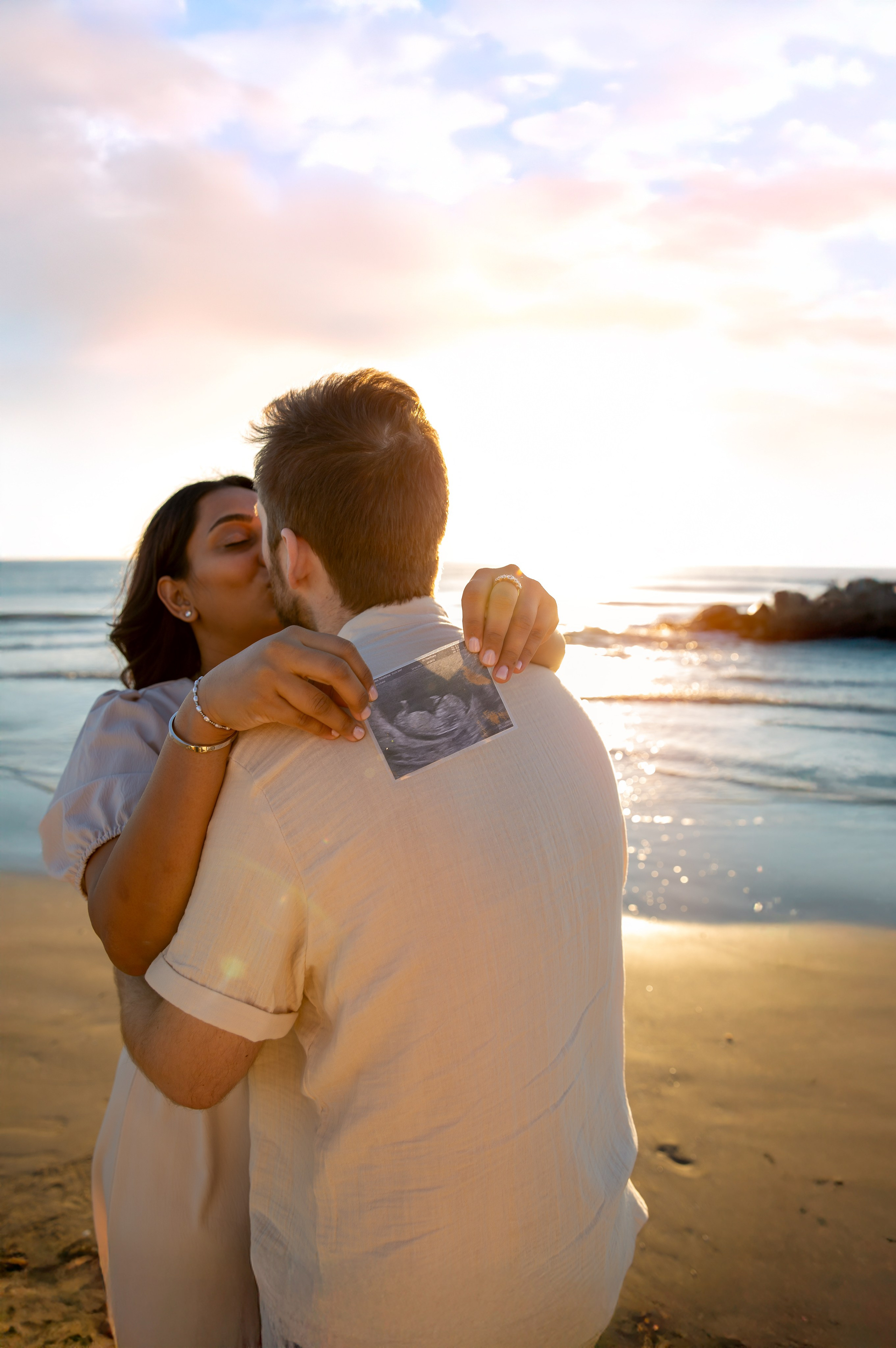 Sesión de fotos de maternidad romántica en la playa de Valencia, España — pareja besándose al atardecer mientras sostienen una ecografía, capturando de forma hermosa la alegría y la ilusión del embarazo. Inspiración perfecta para sesiones de maternidad en Valencia y en la costa española.