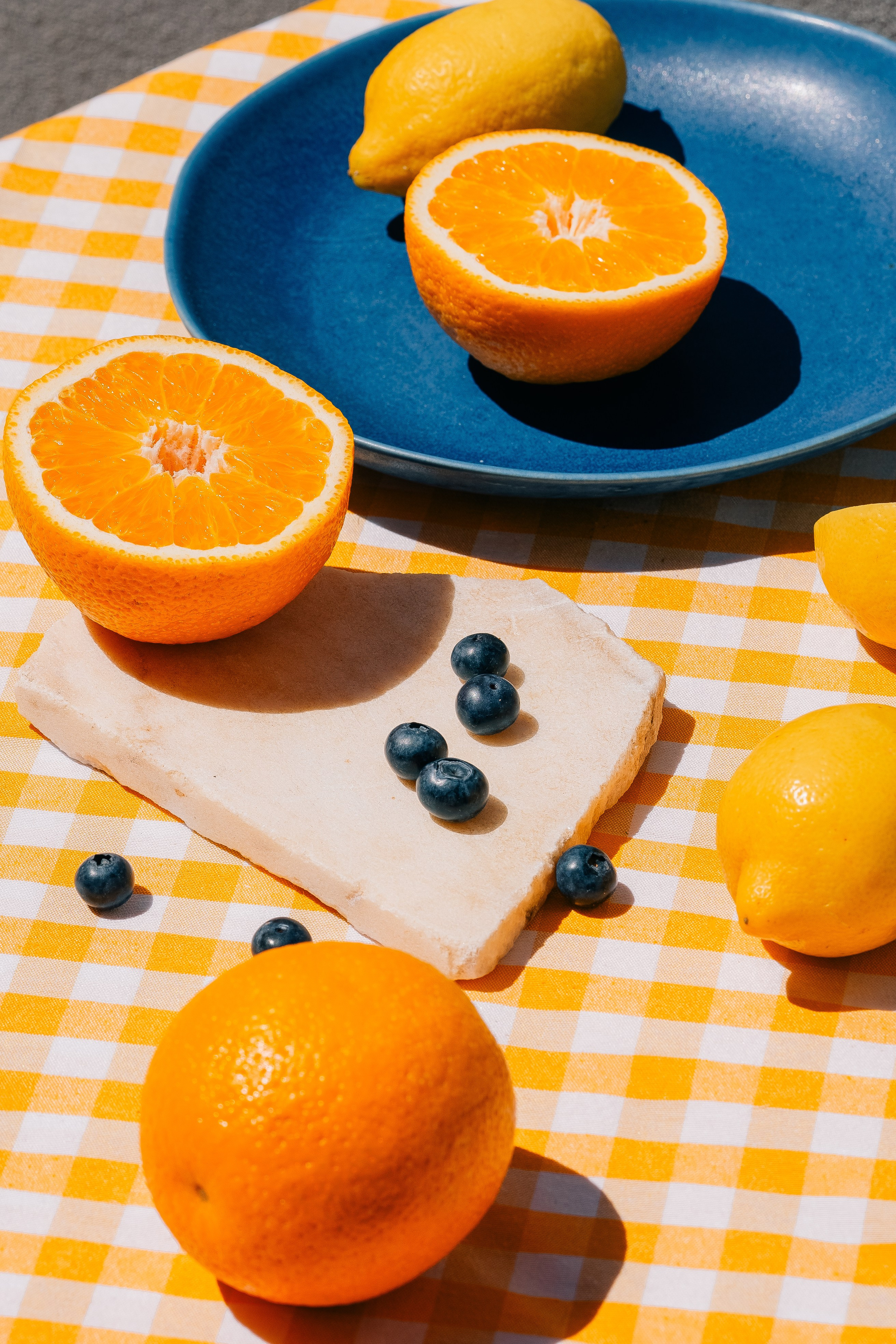 Свежие цитрусовые и черника на клетчатой ​​скатерти. Яркие цитрусовые и черника, разложенные на желто-белой клетчатой ​​скатерти в лучах солнца.