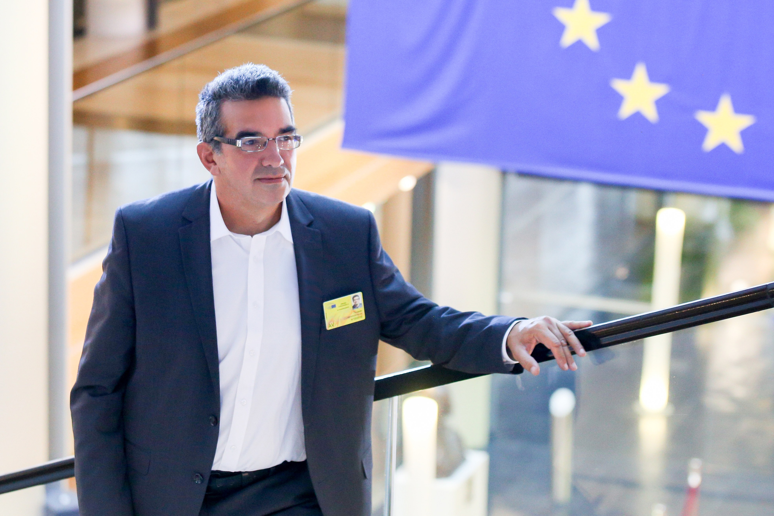 Panagiotis Lymperopoulos, a professional in a dark suit and glasses, poses for a portrait while leaning on a railing inside the modern European Parliament building in Strasbourg, with the European Union flag visible behind him.