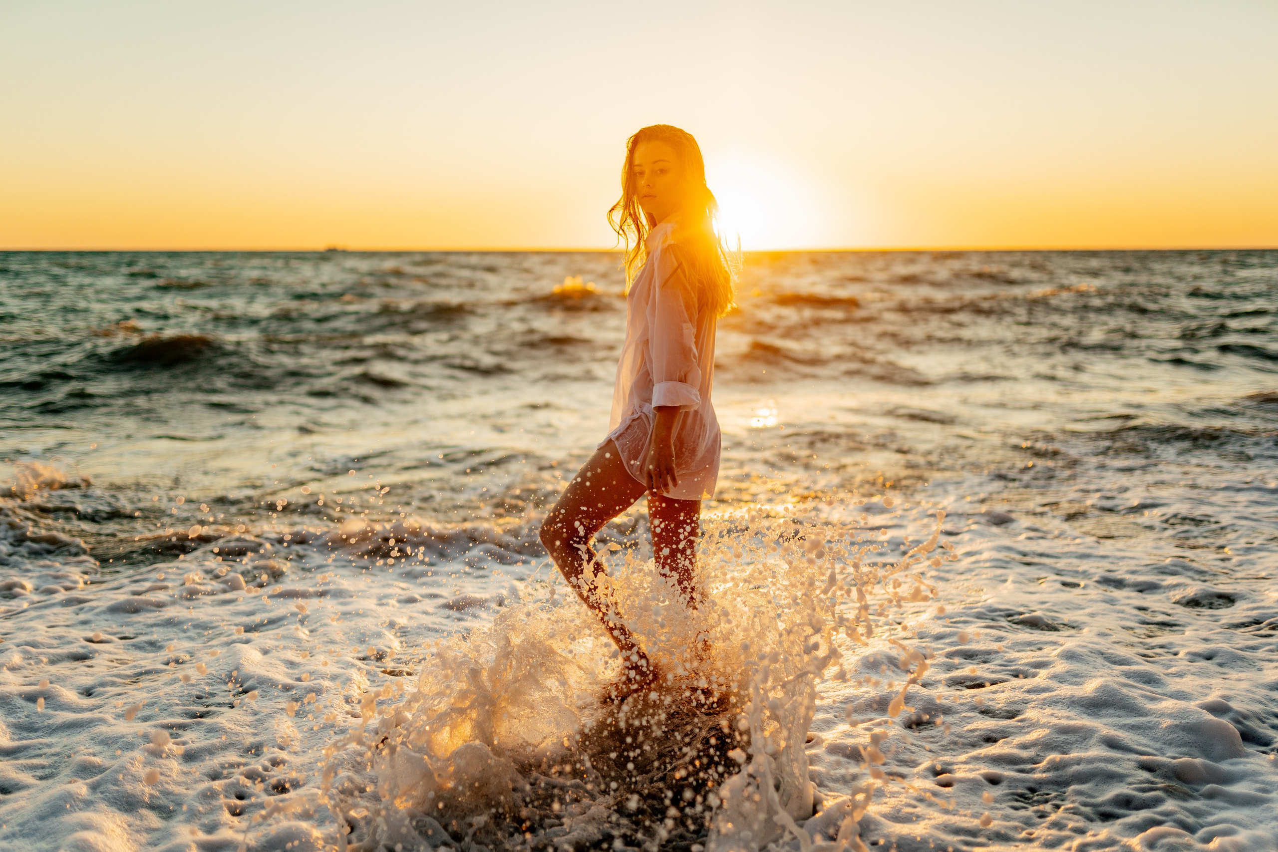 The model, in a white bikini and sheer cover-up, enjoys the waves at sunset. The combination of the setting sun and the ocean waves creates a picturesque and serene moment.