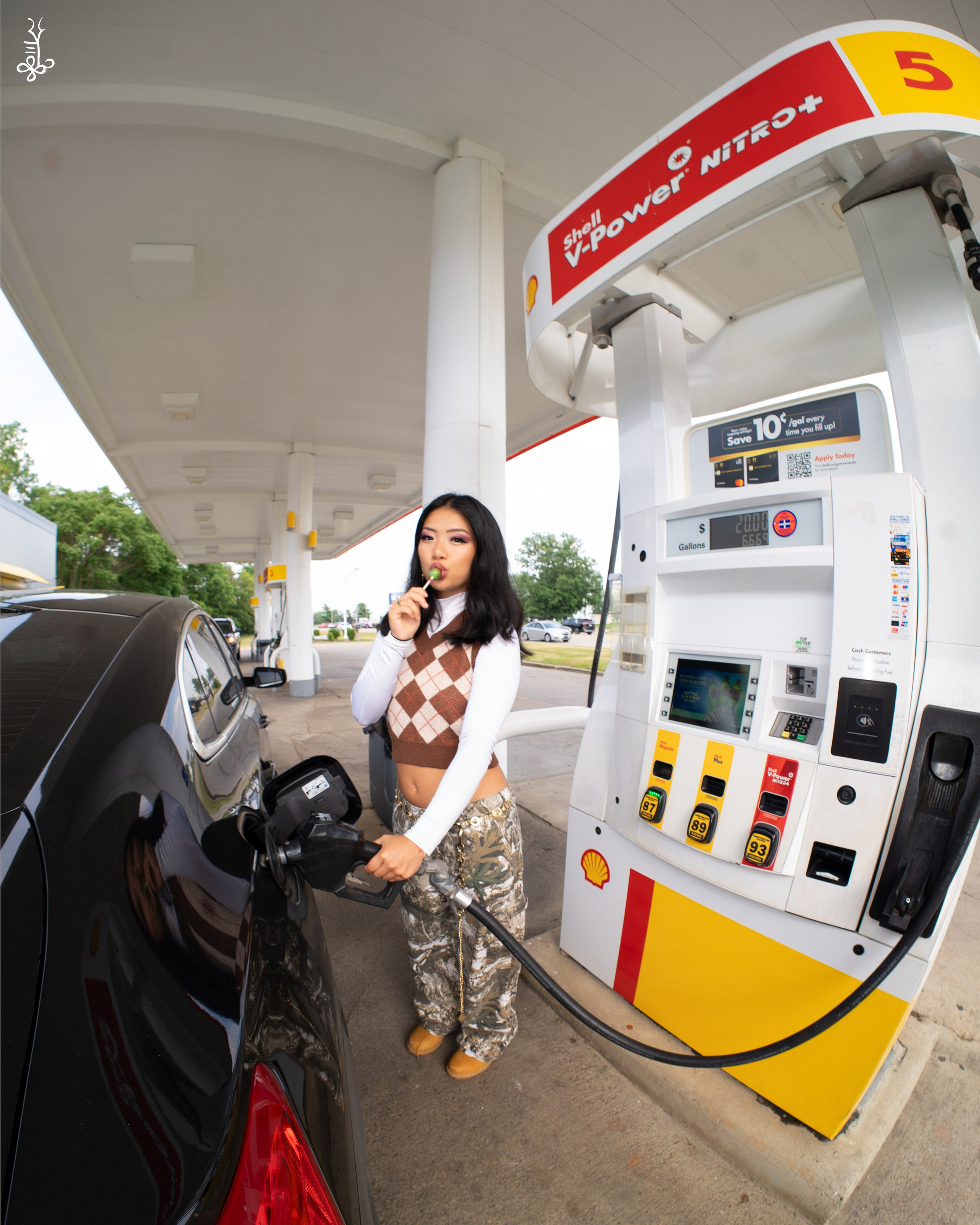 Model pumping gas while standing next to Shell station pump in streetwear outfit
