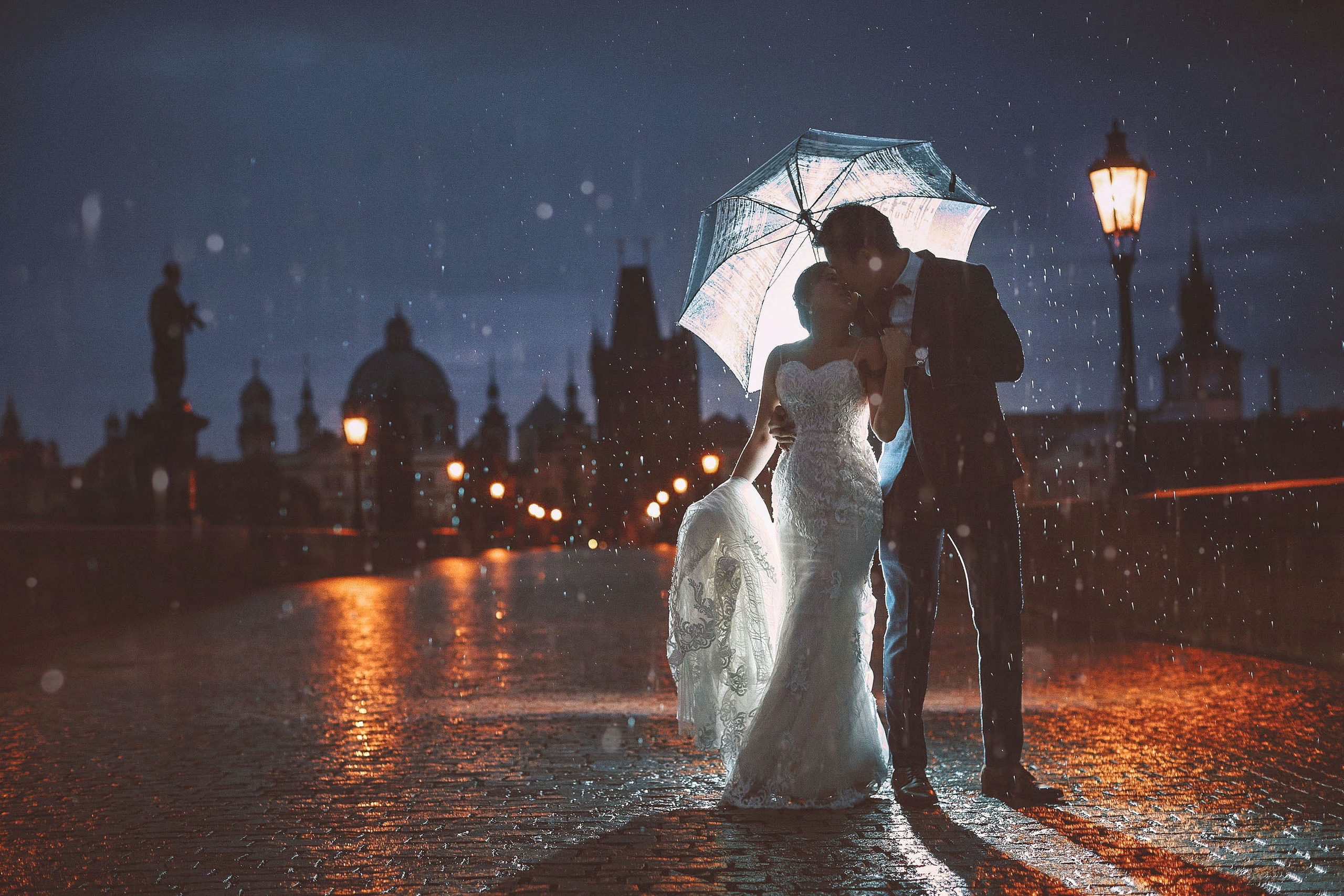 Couple in wedding clothes sharing an umbrella in the rain on Charles Bridge at night for a pre-wedding shoot in Prague.