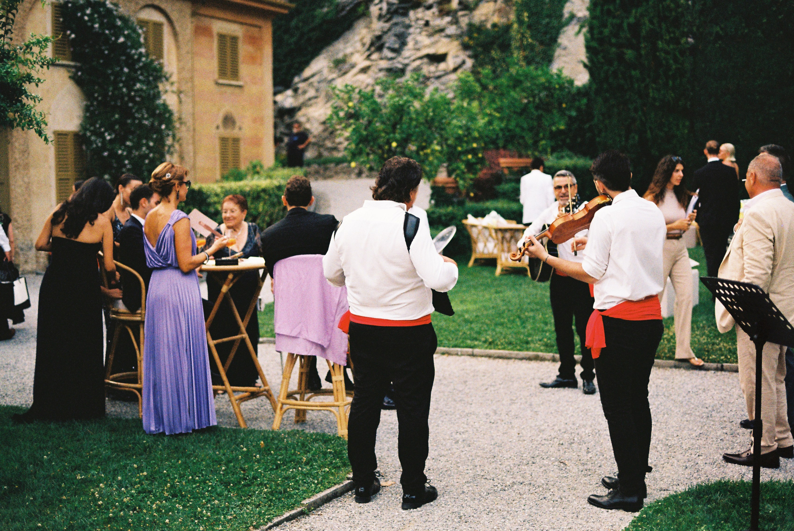Guests gather around musicians playing guitar and violin during outdoor wedding reception.