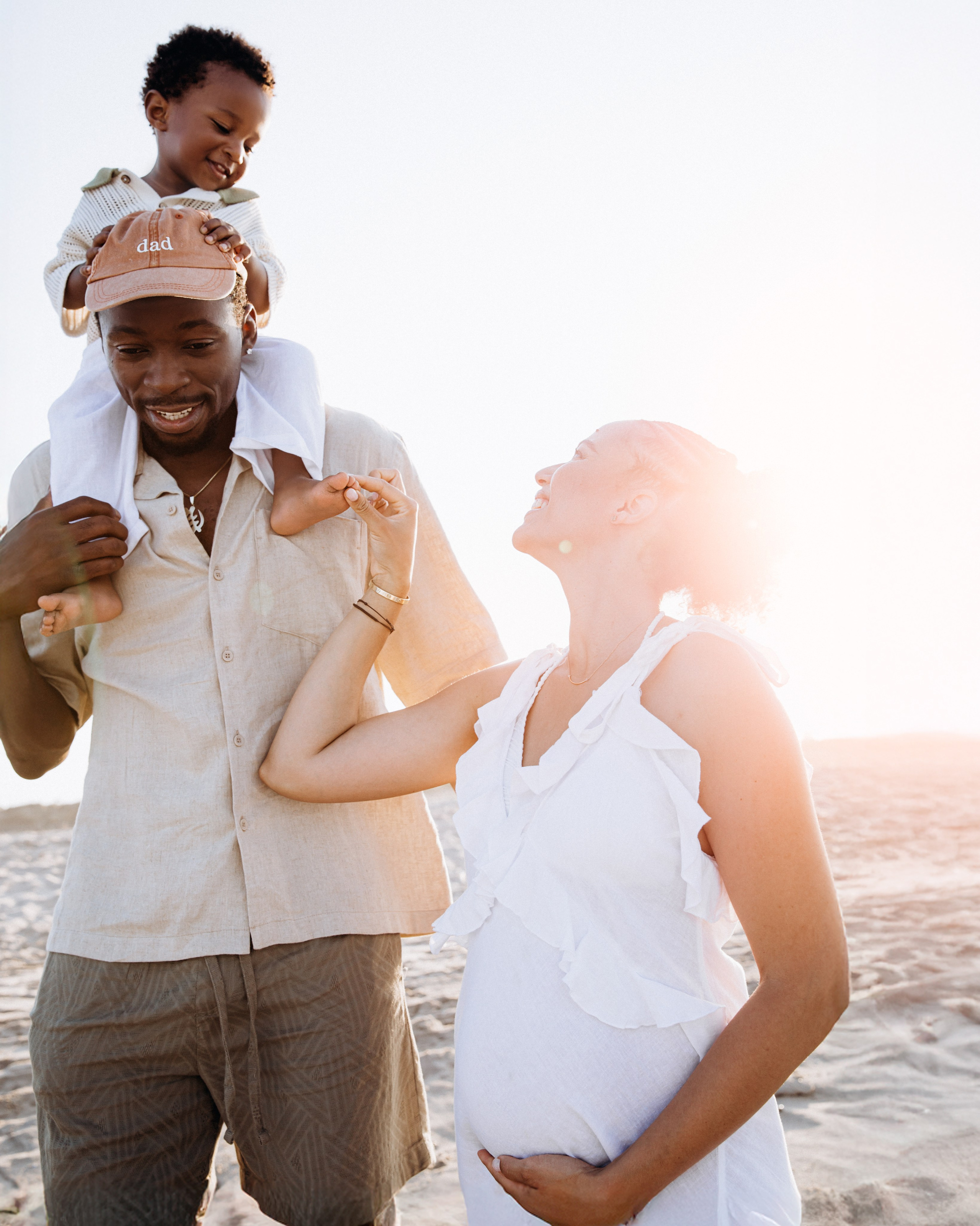 Momento familiar divertido capturado en la playa de Alicante, España — padre sonriente con su hijo sobre los hombros y madre embarazada a su lado. Ideal para sesiones familiares naturales y espontáneas en Alicante y la costa española.