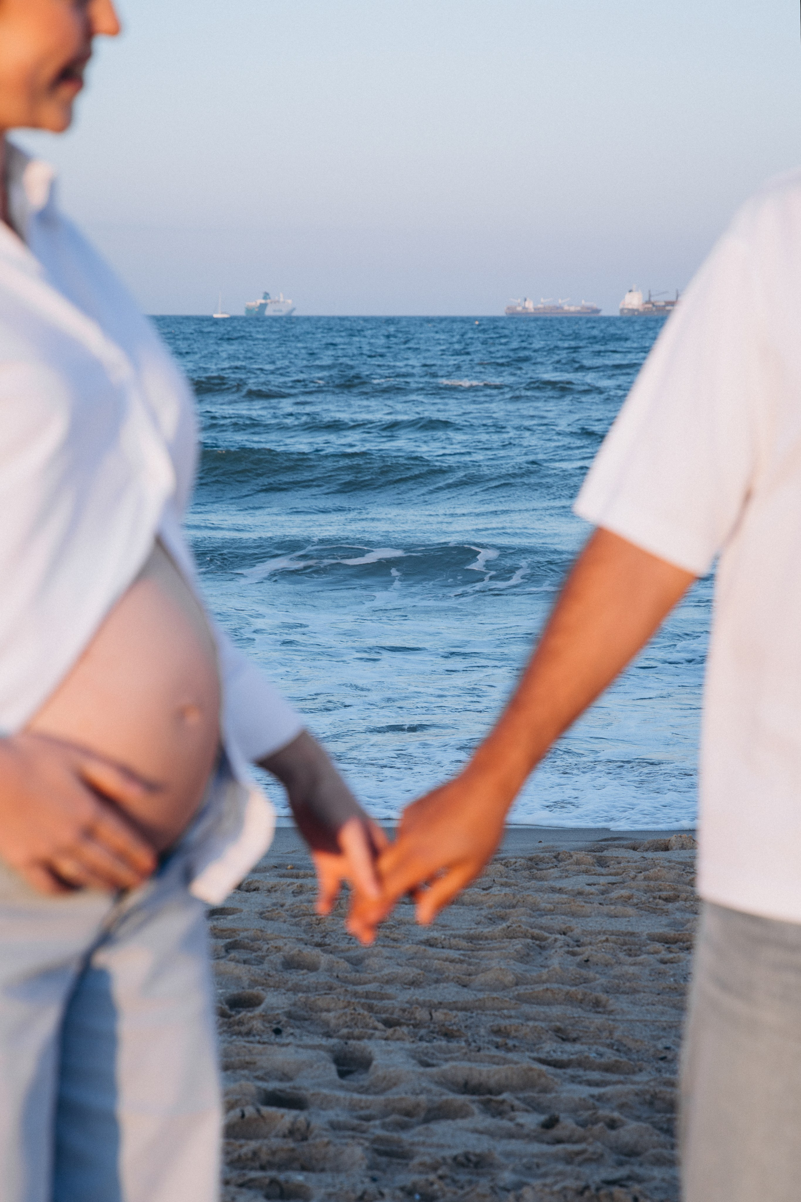 Sesión de fotos de embarazo en Valencia, España, con un primer plano de una pareja embarazada cogida de la mano en la playa con la barriga visible, capturada con luz natural suave junto al mar — ideal para sesiones maternity en Valencia y en toda España.