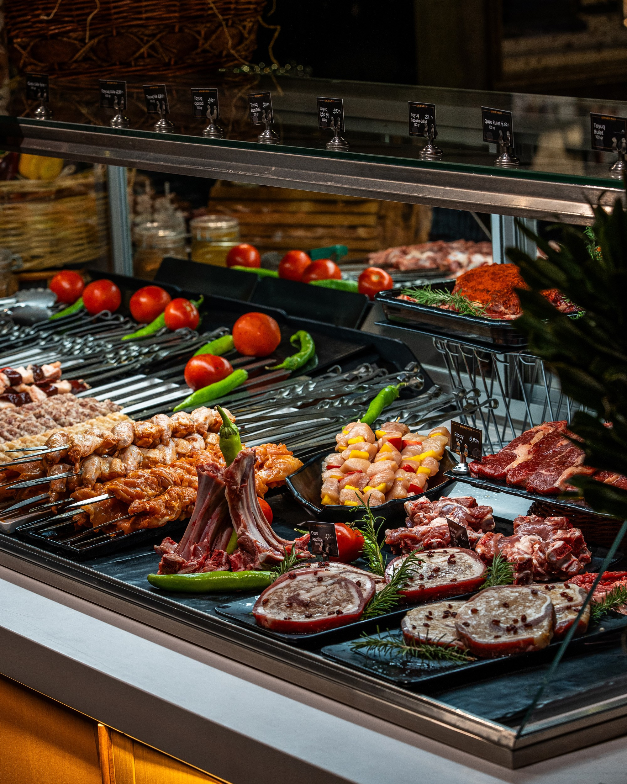 Soyuducu vitrin içində nümayiş olunan müxtəlif növ təzə ət məhsulları, o cümlədən şişlər, quzu qabırğaları, döş əti rulonları və steyklər, pomidor və bibərlə bəzədilmiş.