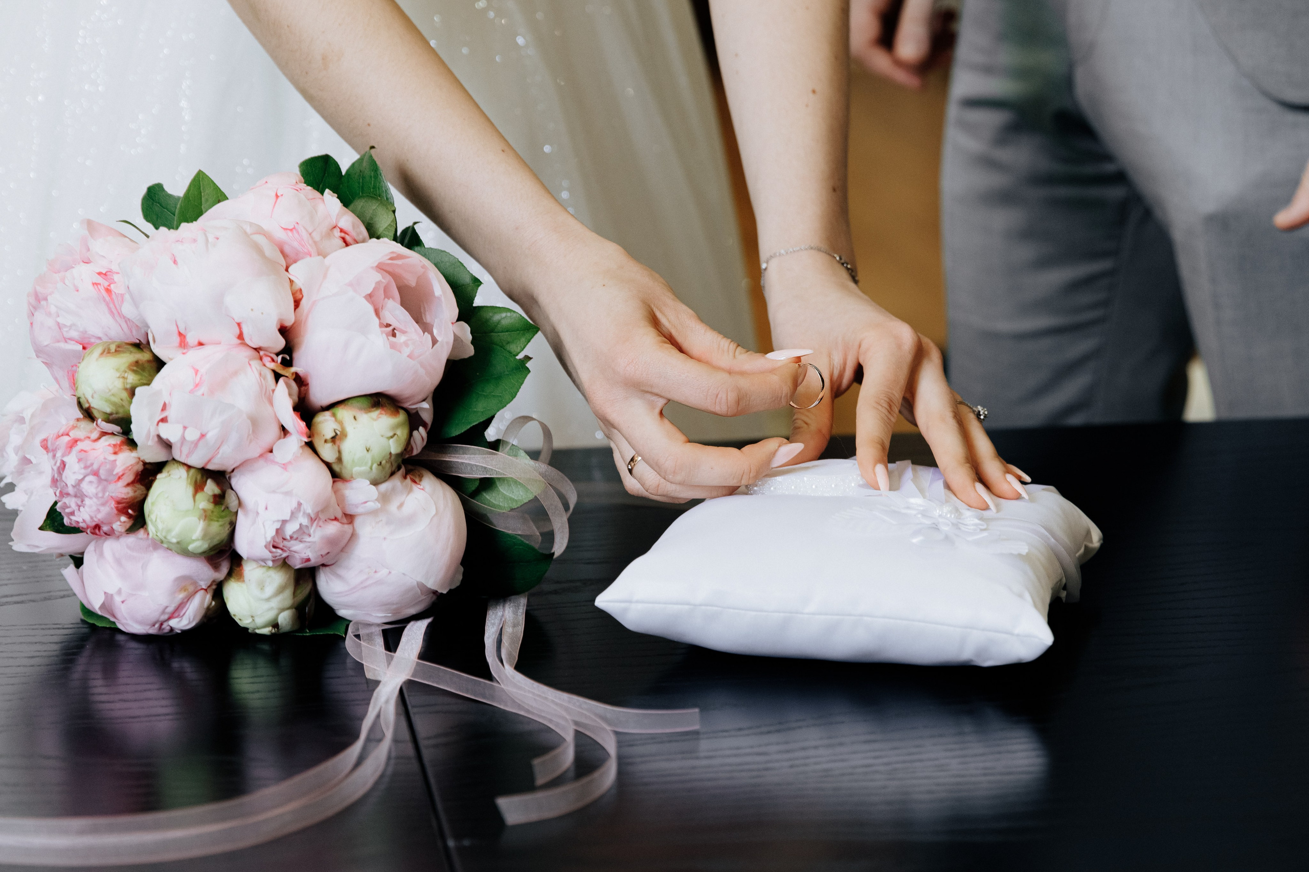 Die Braut nimmt den Ehering von der Kissen, während daneben der Brautstrauß aus Pfingstrosen liegt.