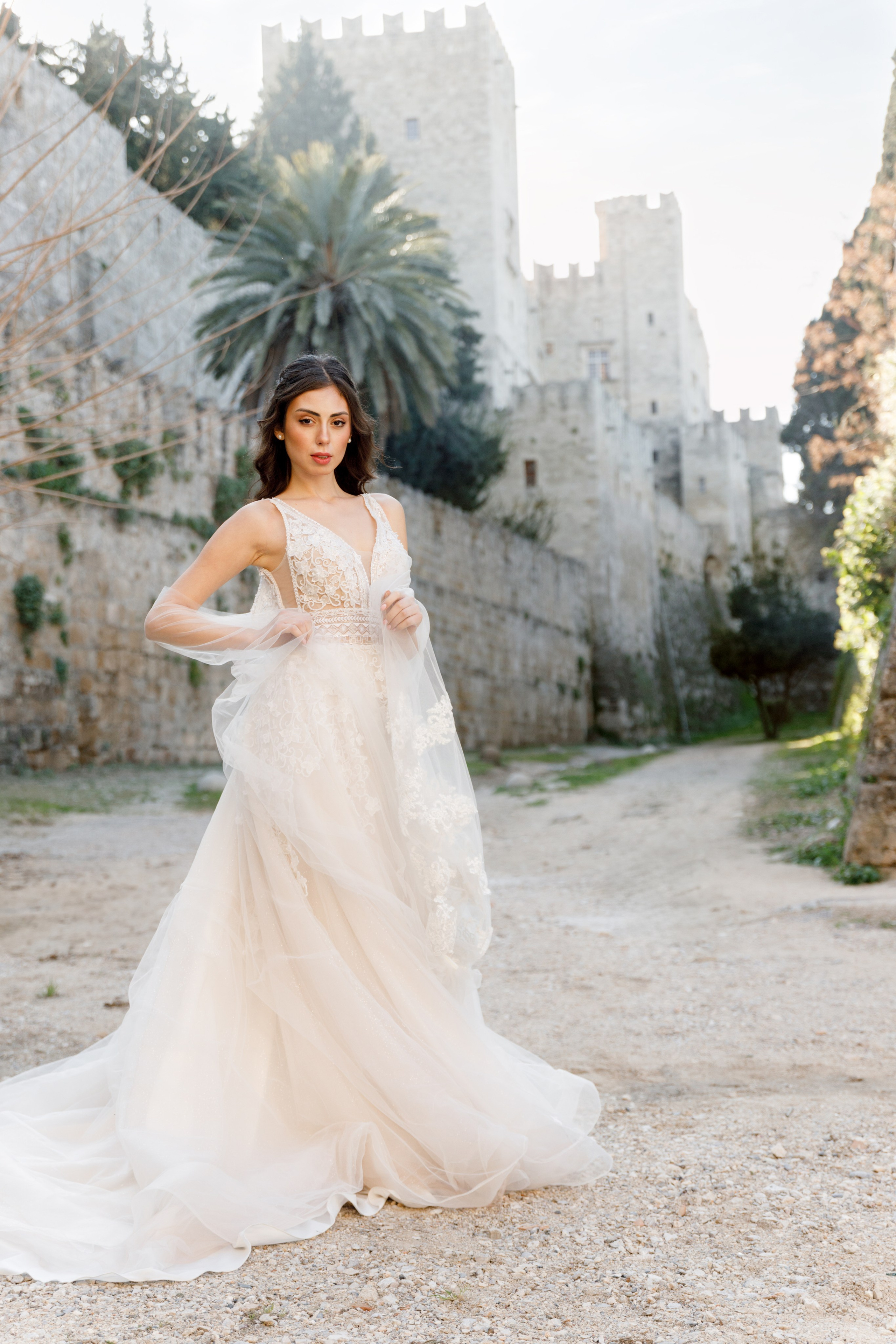 A stunning bride gazes thoughtfully in the enchanting alleys of Rhodes' Old Town, her flowing wedding dress complementing the rustic charm of the cobblestone streets and ancient architecture. The editorial-style portrait captures her poise and the romantic atmosphere of the medieval surroundings, bathed in warm, golden light.