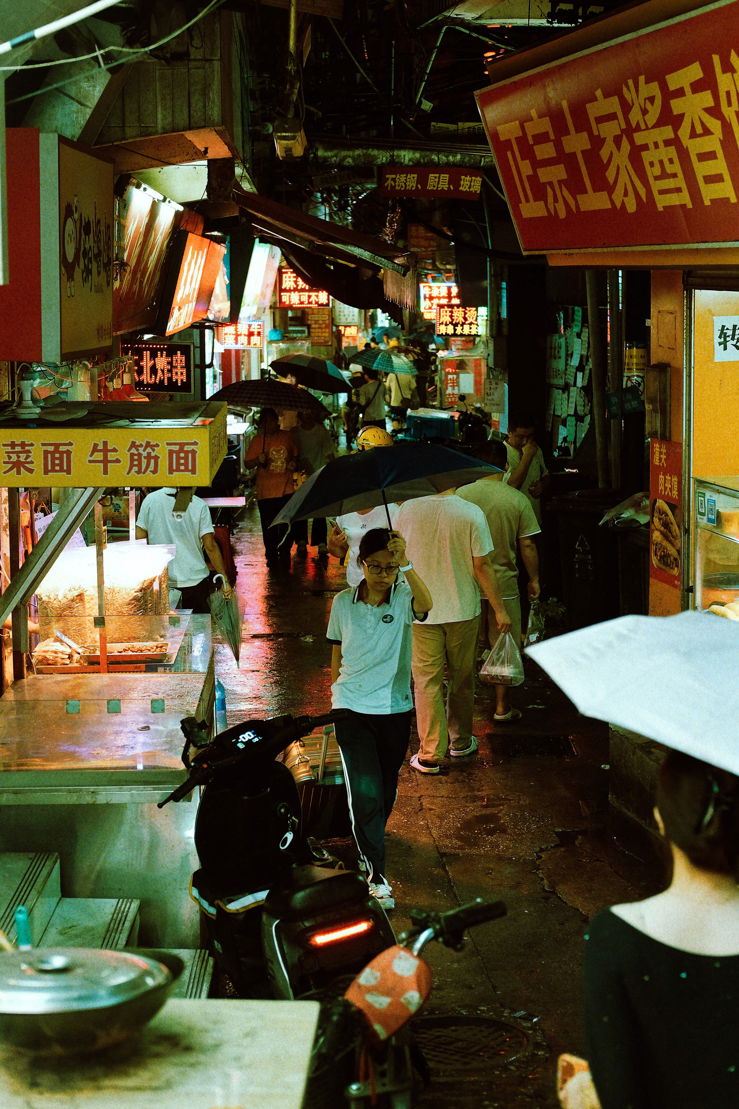 Rainy evening street scene in Guangzhou with pedestrians carrying umbrellas through a neon-lit market alley, photographed with a Fujifilm X-Pro3 and finished with a cinematic film-inspired edit.