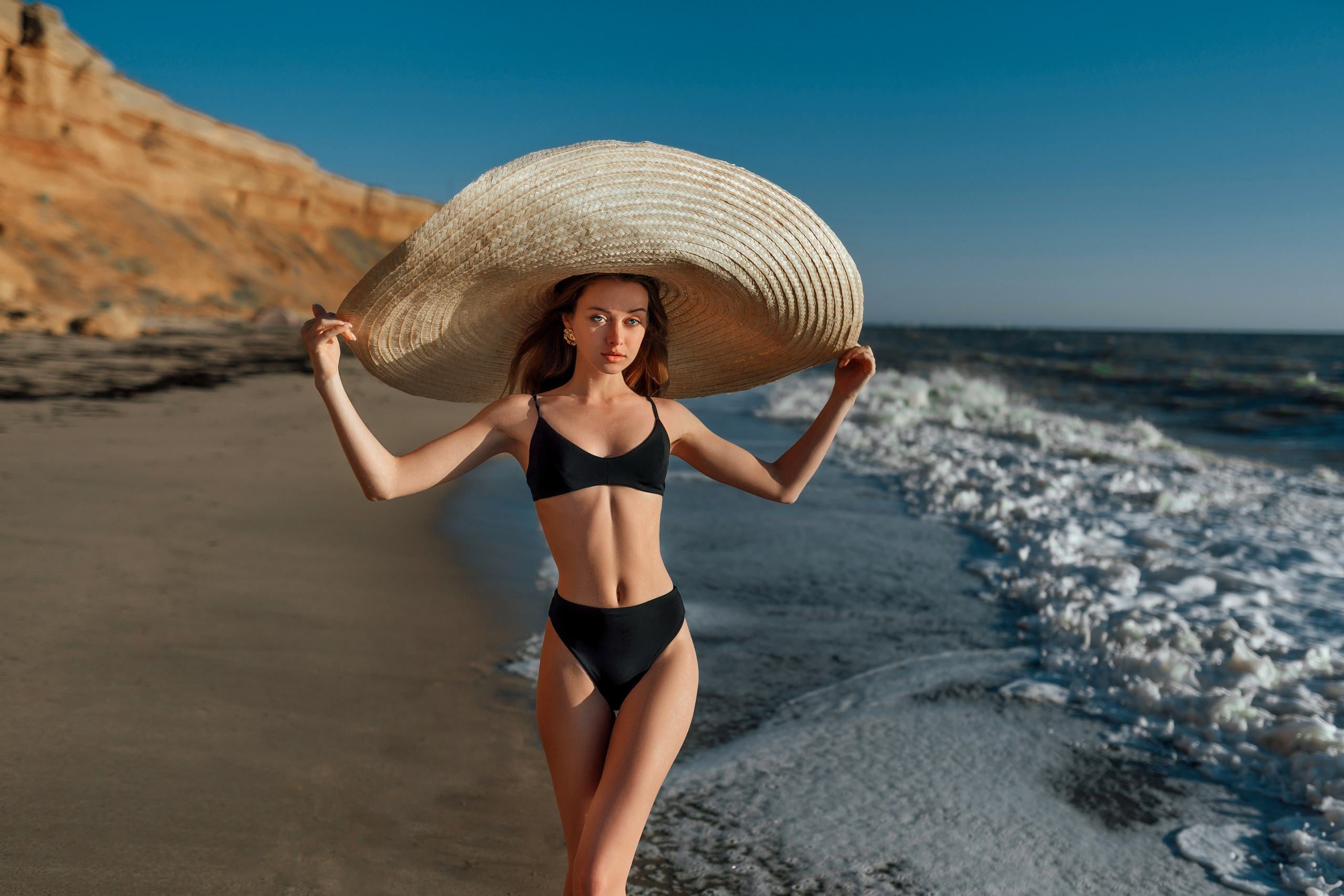 A stunning model showcases beachside elegance in a black bikini, complemented by a wide-brimmed straw hat. The clear blue sky and crashing waves create a perfect backdrop for this fashionable beach scene. Beachside, elegance, black bikini, straw hat, fashion, ocean backdrop