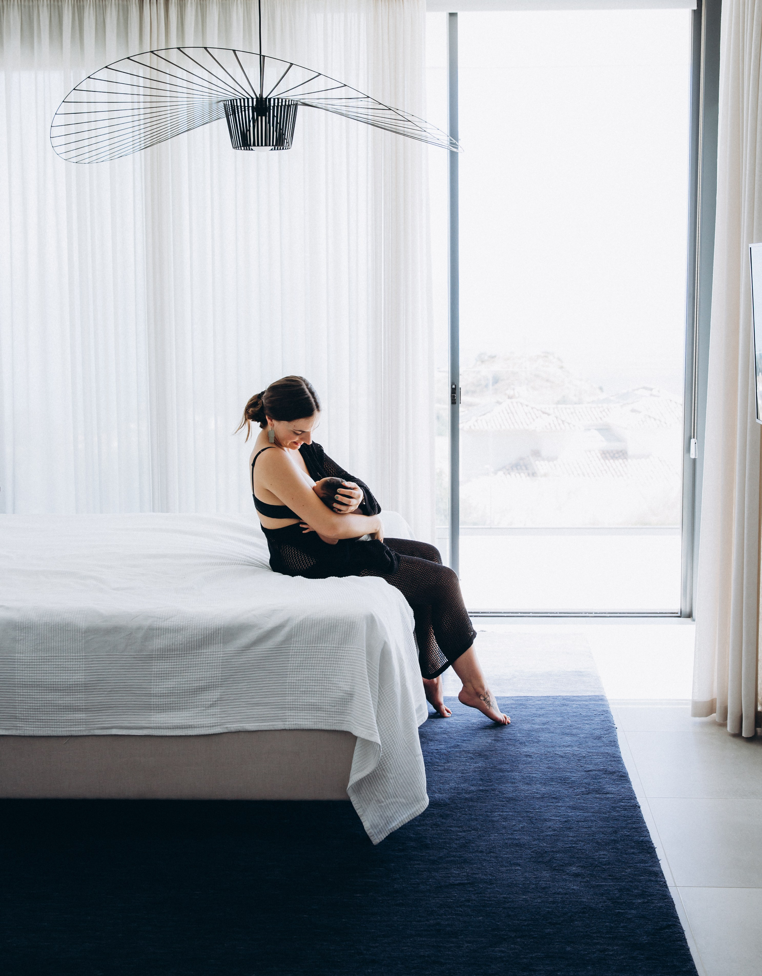 Fotografía familiar minimalista en Alicante, España — madre en calma acunando a su recién nacido en un dormitorio moderno y luminoso con luz natural suave. Perfecto para quienes buscan sesiones íntimas y elegantes.
