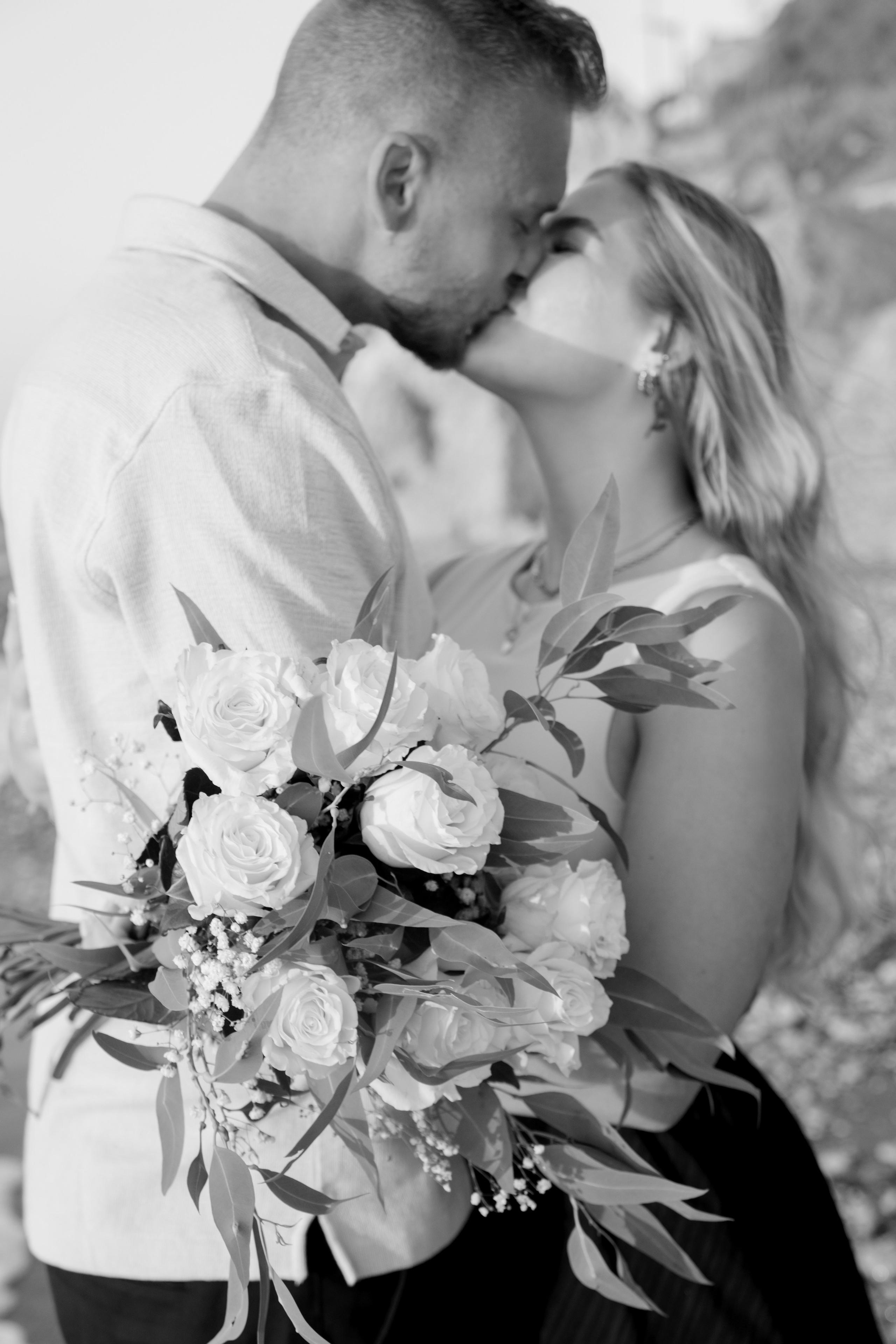 "Ein glücklicher Moment am Kato Petres: Ein Paar schaut sich verliebt an, umgeben von den markanten, ockerfarbenen Felsformationen und dem Rauschen des Meeres. In ihren Händen halten sie einen Strauß strahlend weißer Rosen, der einen eleganten Kontrast zur rauen Schönheit der steinigen Küste bildet, während das warme Abendlicht auf den Blüten und den Wellen glitzert."