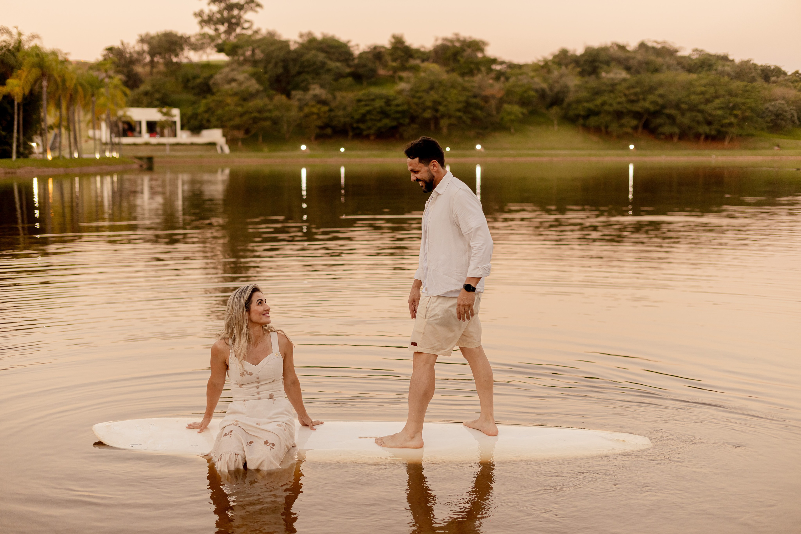 Casal sentado em prancha de SUP no meio do lago ao entardecer, com reflexo das palmeiras na água no Resort das Oliveiras