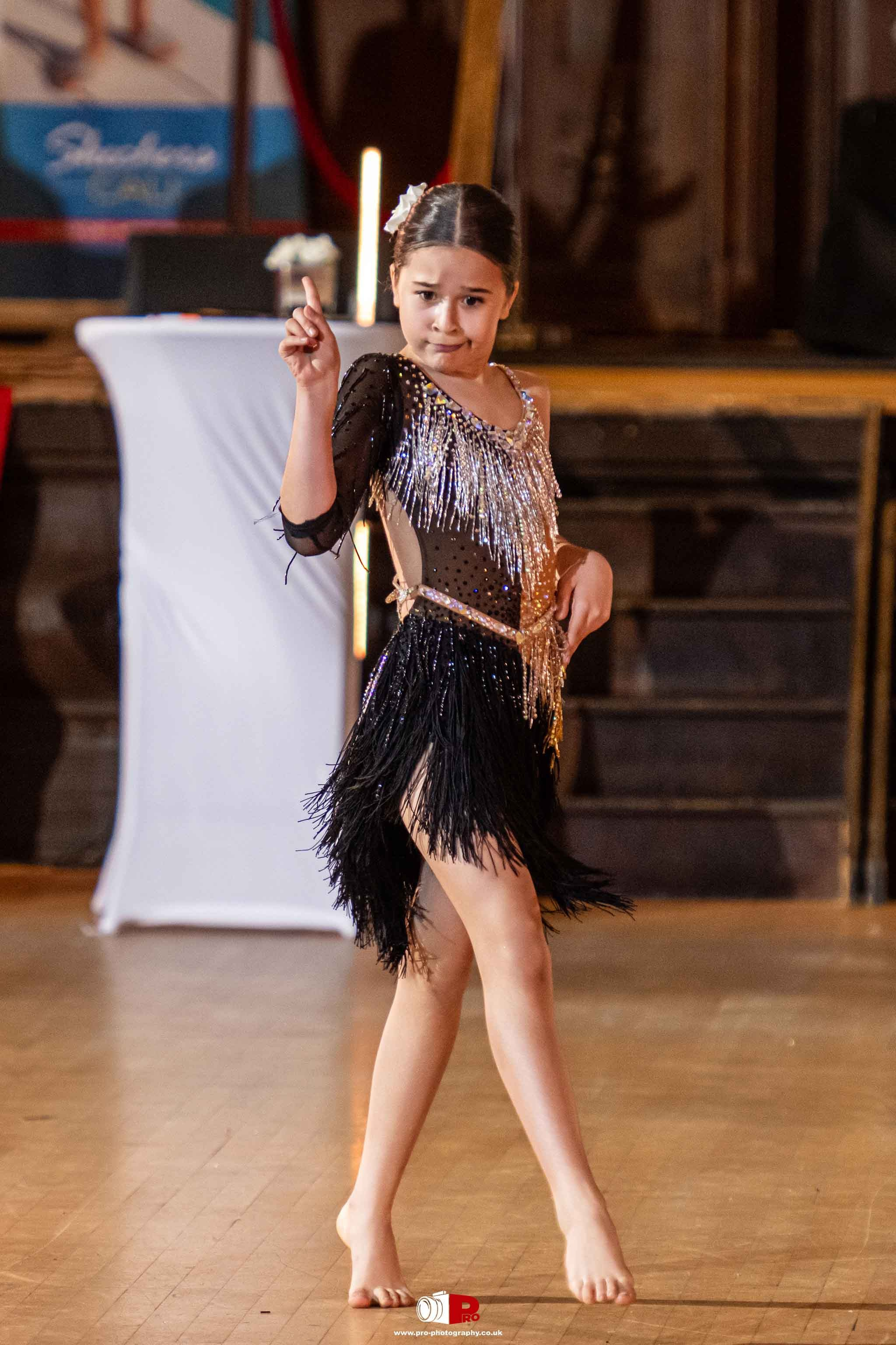 A young dancer in a sparkly black and silver costume striking a confident pose on a polished wooden dance floor.