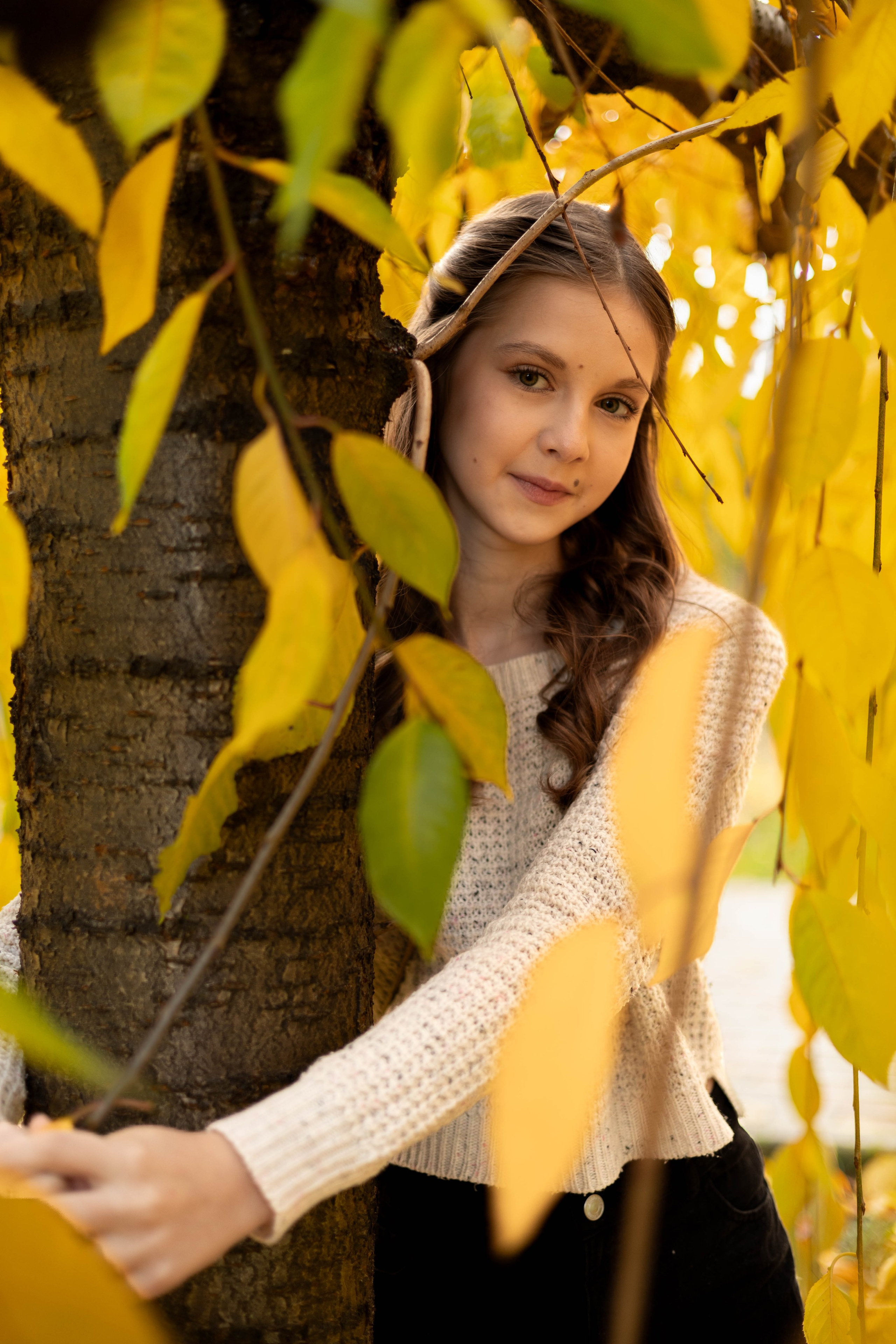 Крупный план лица девушки с лёгкой улыбкой на фоне осенних деревьев.