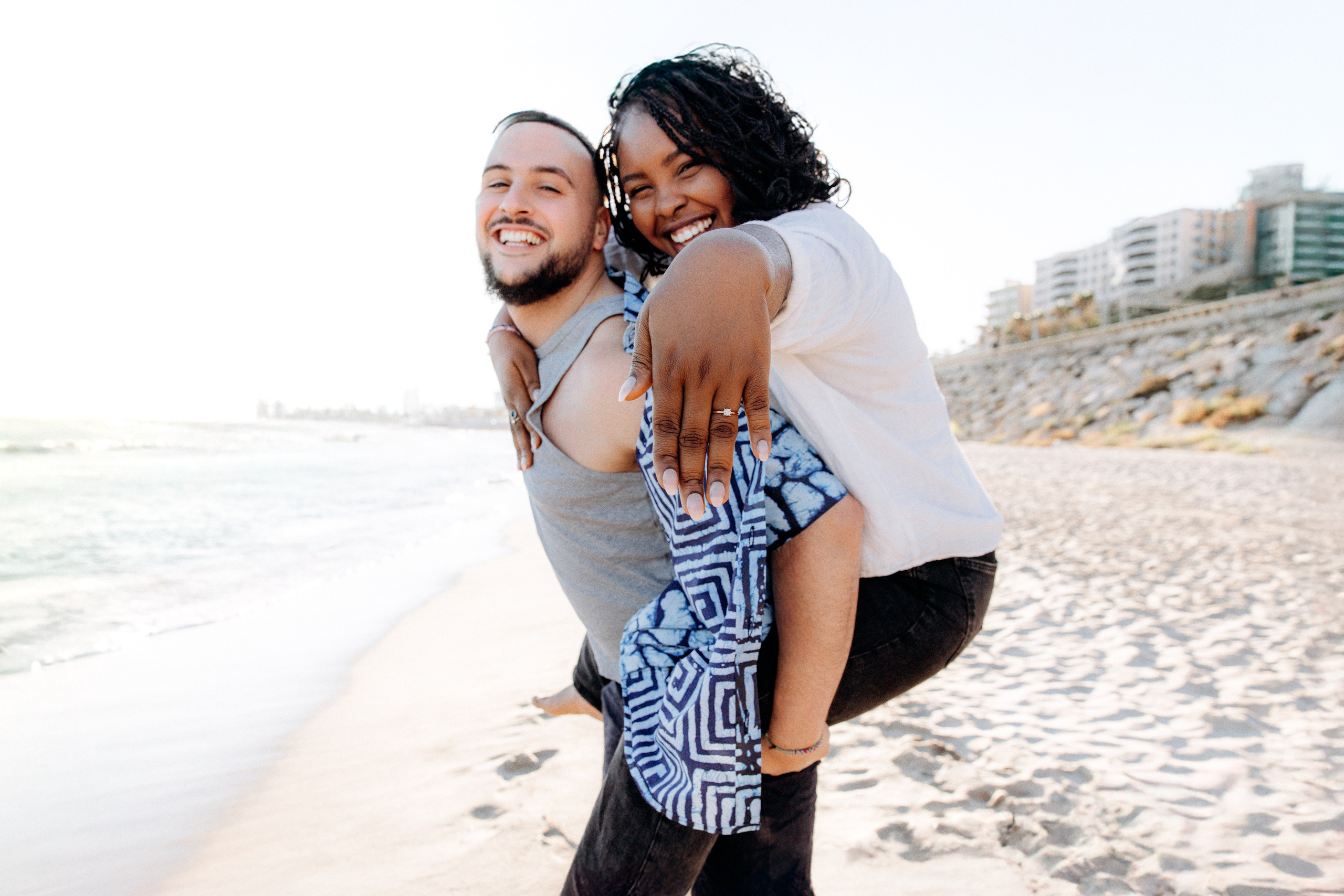 Sesión de fotos divertida en la playa de Valencia, España — una mujer alegre muestra su anillo de compromiso mientras va a caballito, capturando risas y romance. Perfecto para quienes buscan sesiones de fotos de compromiso o historia de amor en Valencia y en toda España.