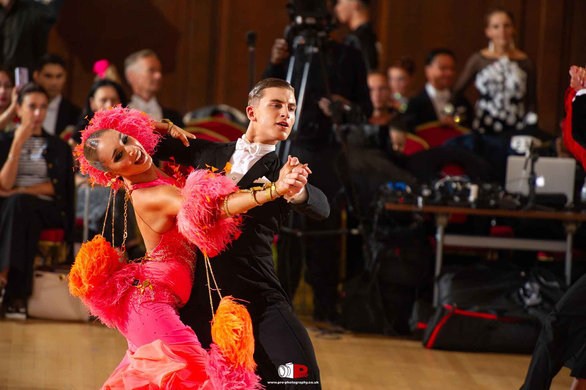 Energetic ballroom dancers in fiery orange and pink costumes showcasing a powerful dance move.