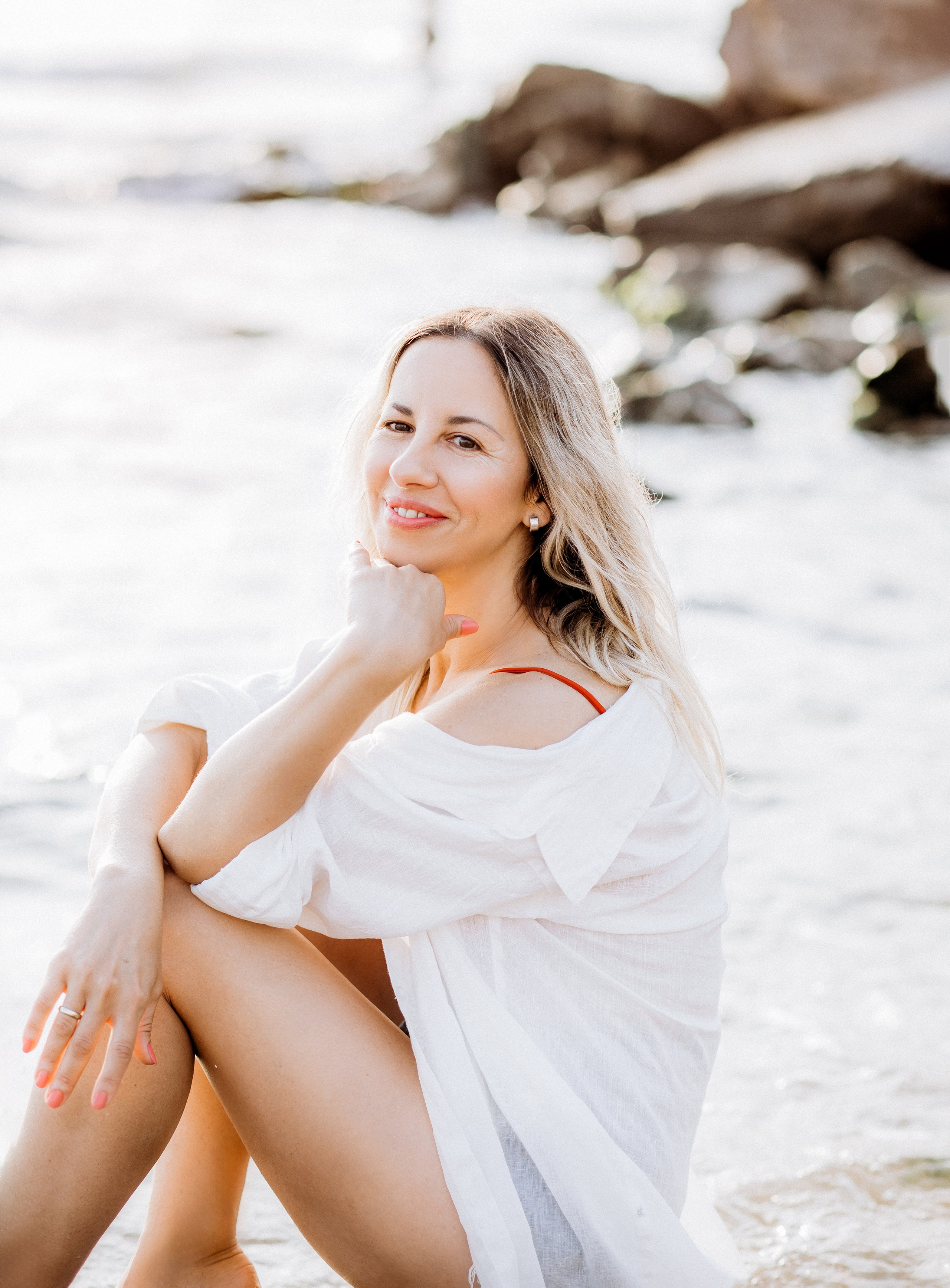 Улыбающаяся женщина сидит у берега в мягком естественном свете, в белой рубашке на тихом пляже в Дении, Испания — расслабленная и элегантная пляжная портретная фотография, передающая природную красоту и прибрежное спокойствие, идеально подходит для портретных фотосессий в Дении и Испании.