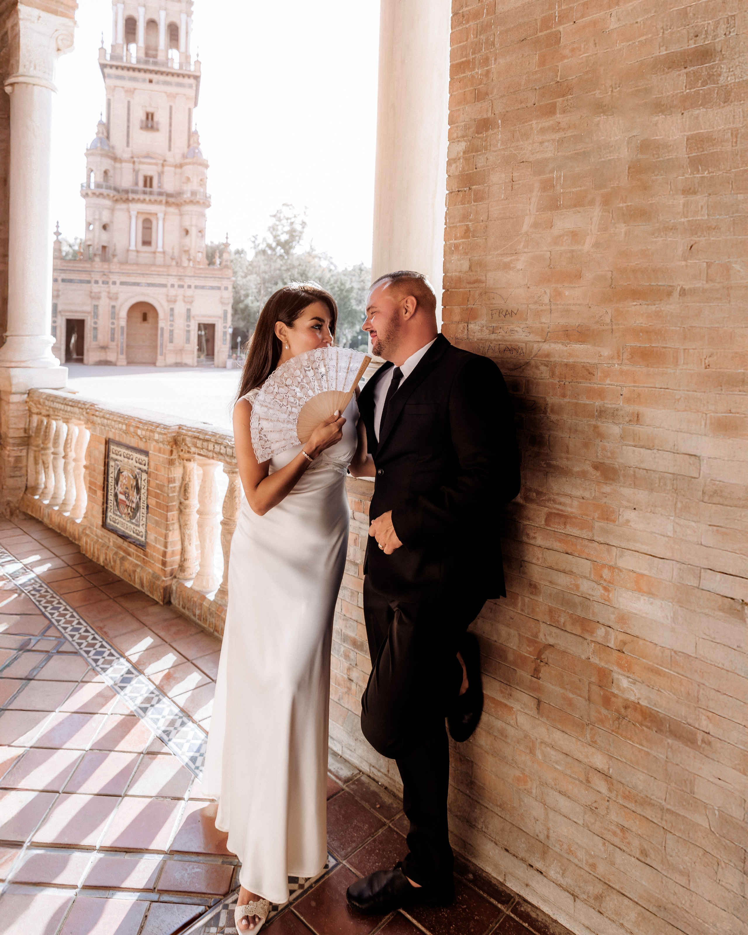 Elegant wedding photo in Sevilla, Spain — the bride playfully holds a traditional Spanish fan while sharing a joyful moment with the groom against the stunning backdrop of Plaza de España. A beautiful example of romantic and stylish wedding photography in Seville and throughout Spain.