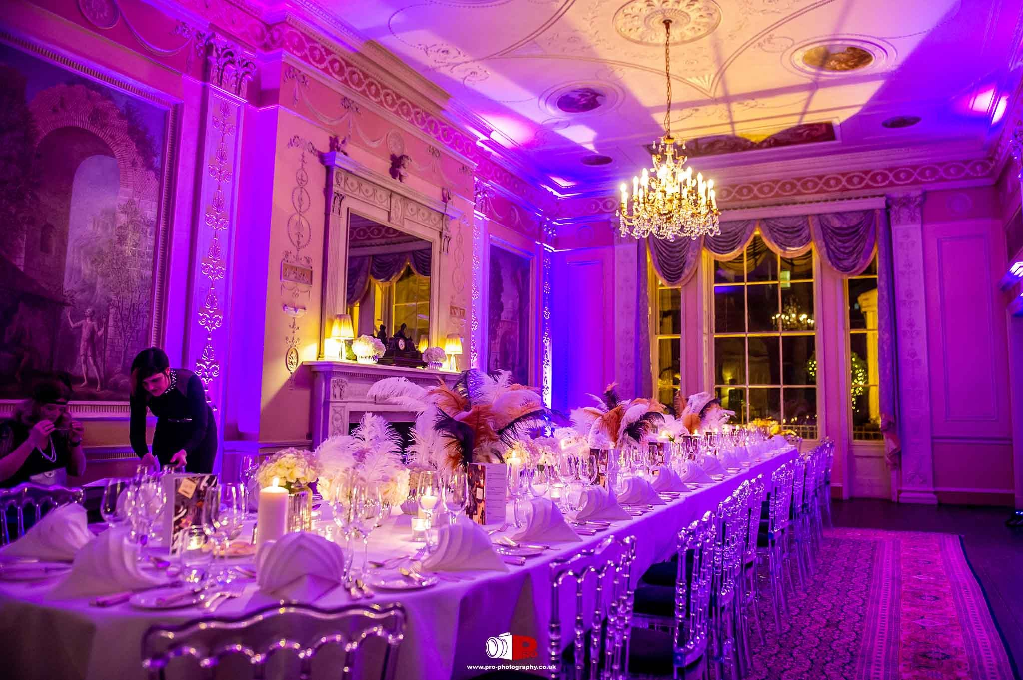 A luxurious dinner table is set up in a grand hall, featuring purple ambient lighting, feather decorations, and crystal-clear glassware.