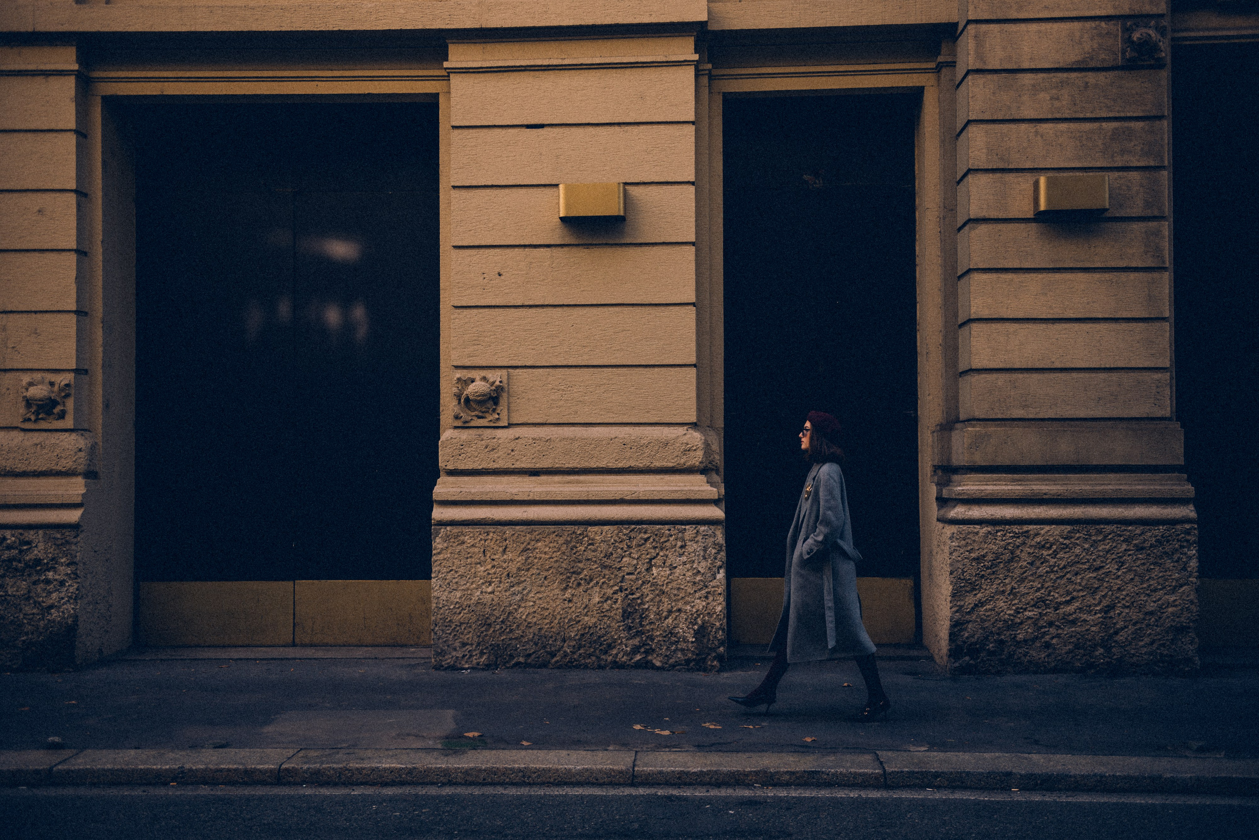 Elegant woman walking along a Milan street in a gray coat and burgundy beret, framed by classic architectural details of the city's buildings