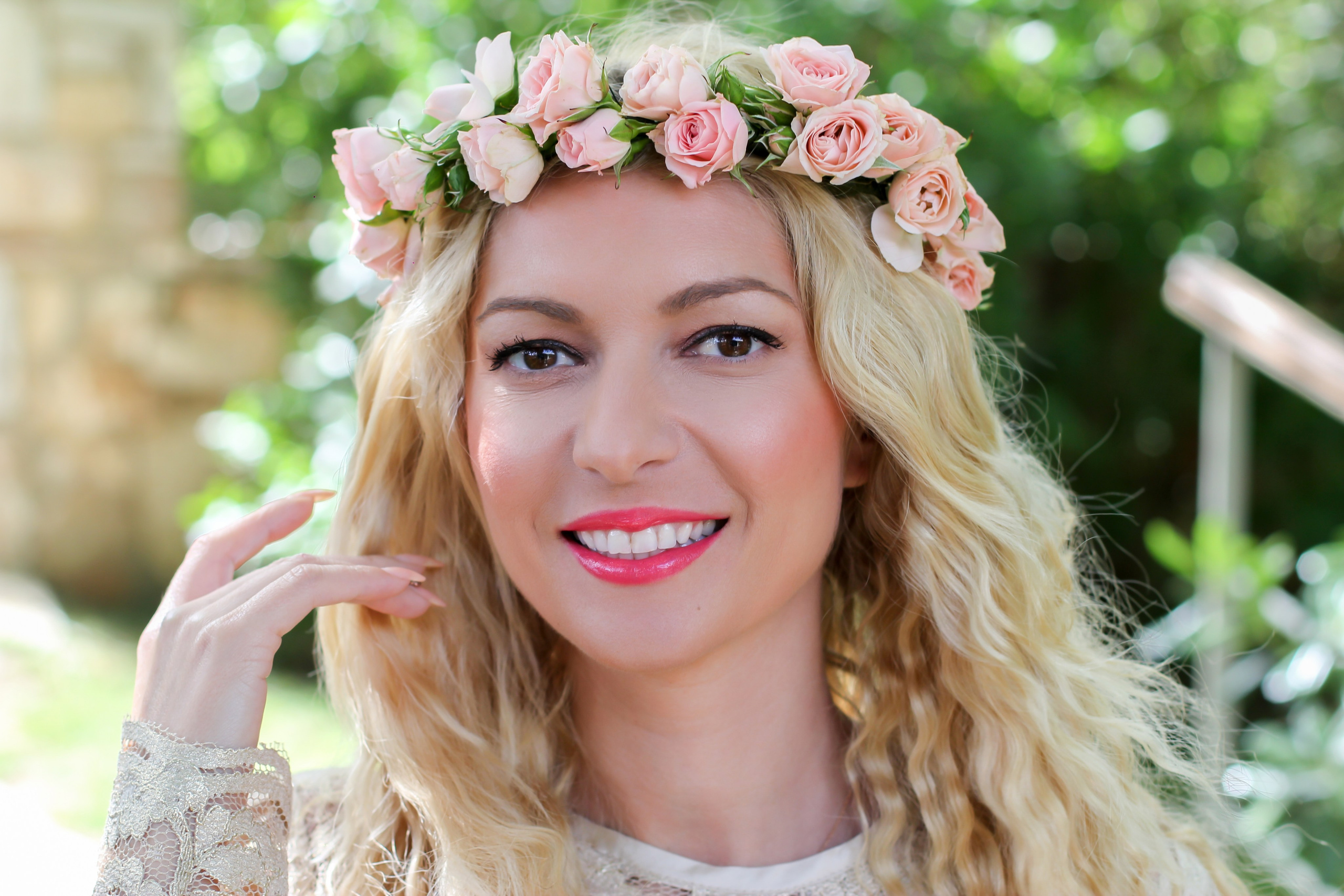 Close-up photo of a smiling blonde woman with wavy hair and bright coral lipstick wearing a delicate pink rose flower crown and a cream-colored lace top.
