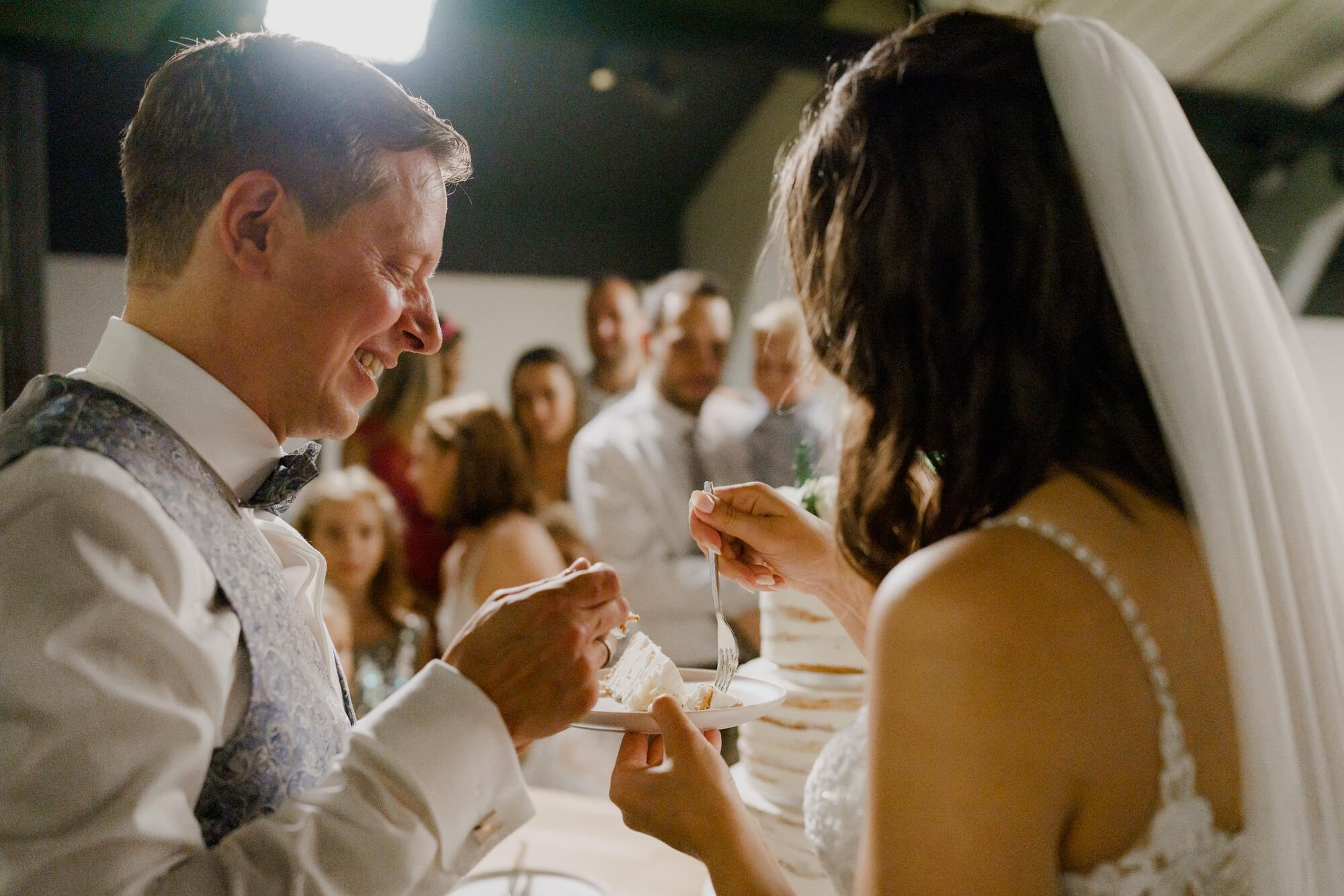 Groom laughing as he and the bride share a piece of wedding cake together, guests watching in the background